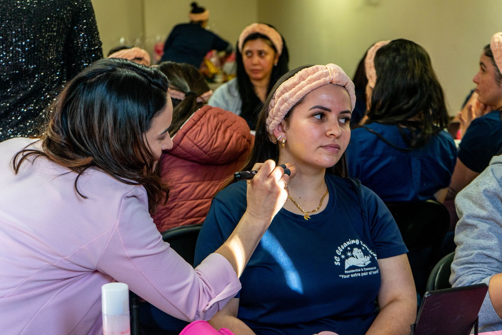 Woman applies makeup to another woman's face. Other women watch in a room.