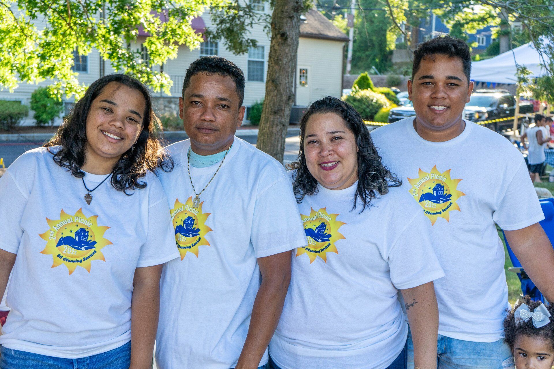 Family of four smiling, wearing matching white t-shirts with a logo, outdoors in daylight.