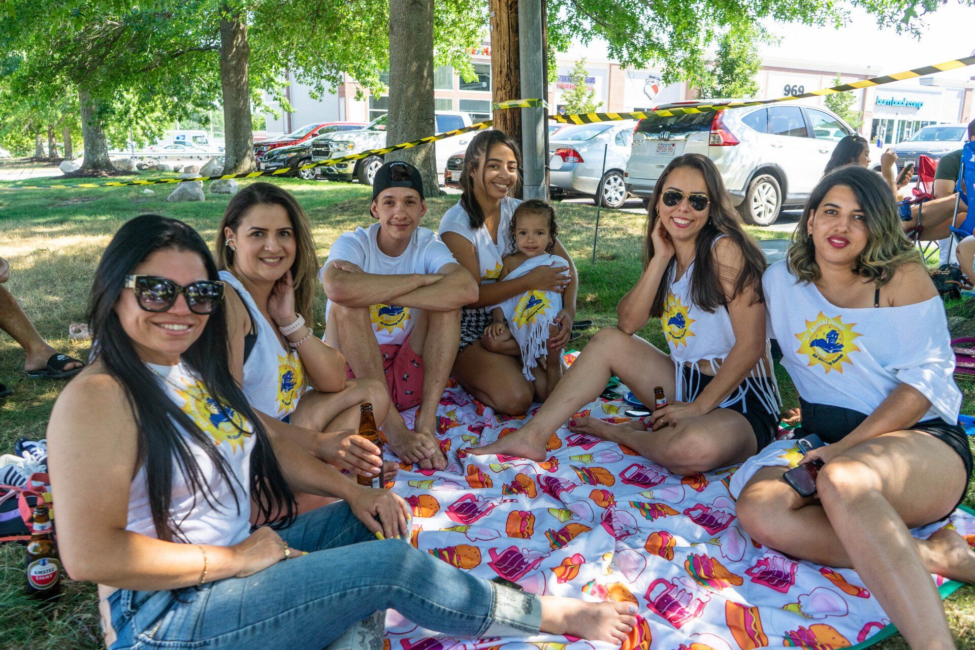 Group of friends smiling on a picnic blanket under trees. Cars and park in the background.