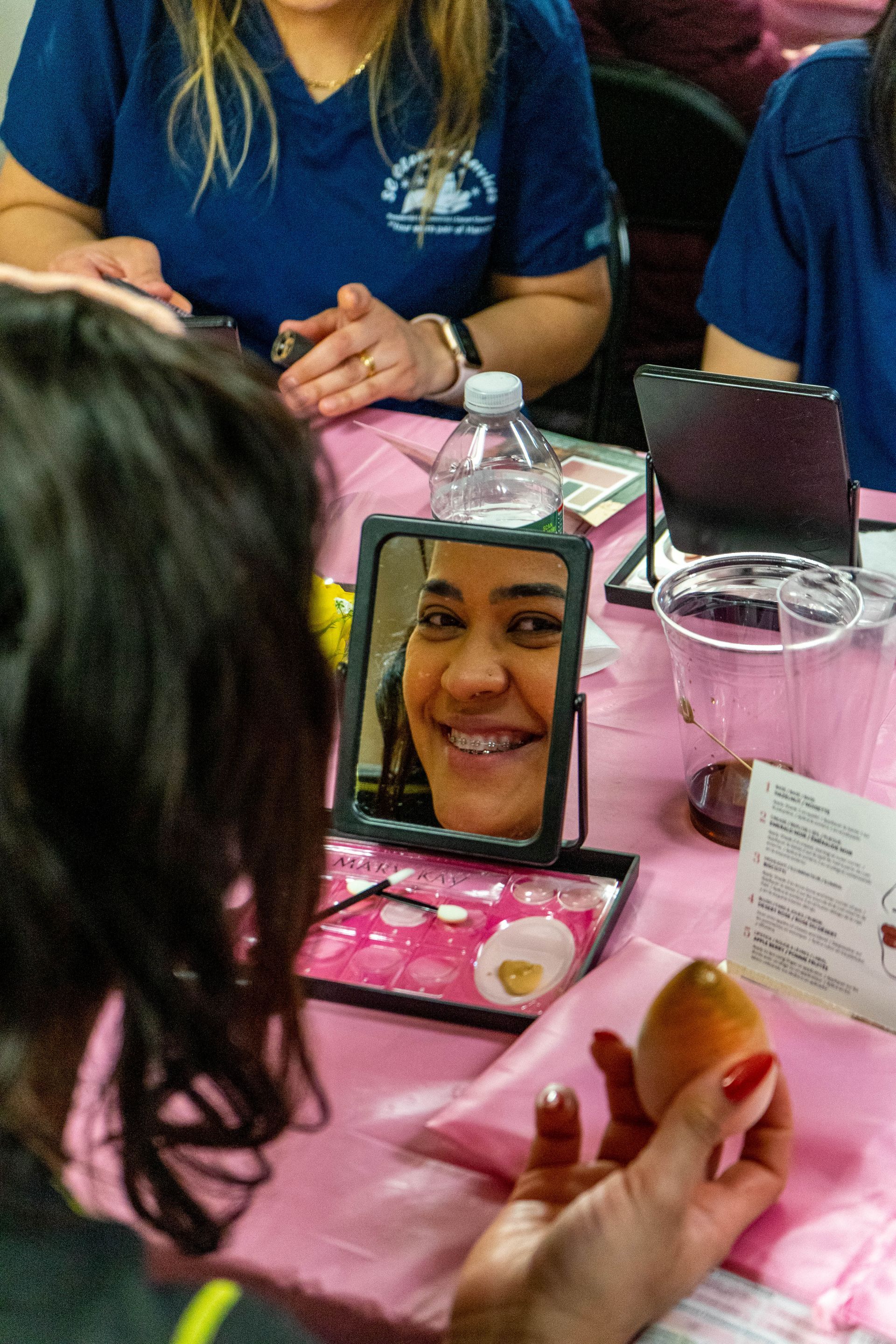 Woman looking in a mirror smiles; a table with beauty supplies is in front of her. Other people are nearby.