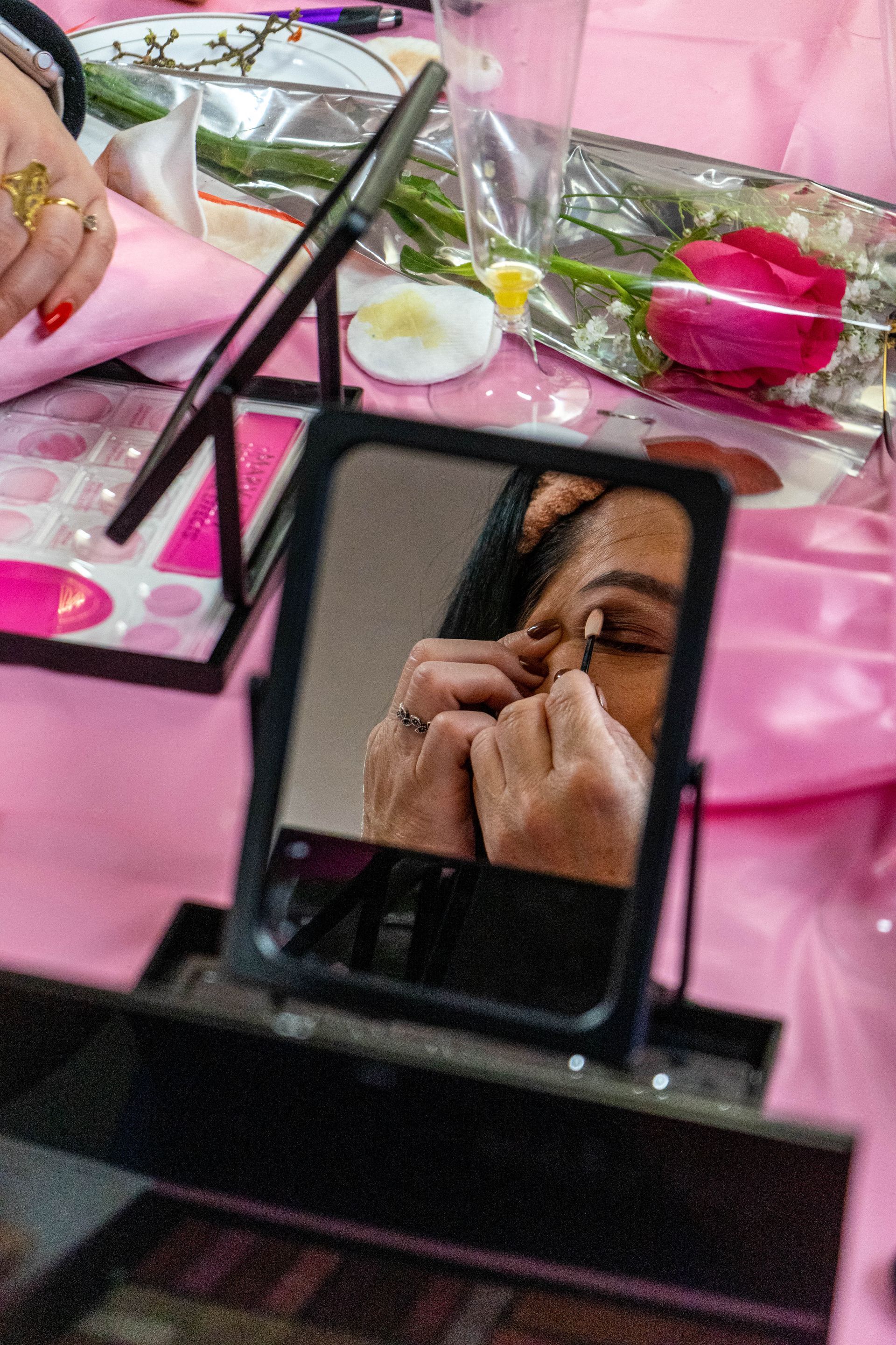 Woman applying eyeshadow in a mirror at a makeup table with pink decor and a rose.