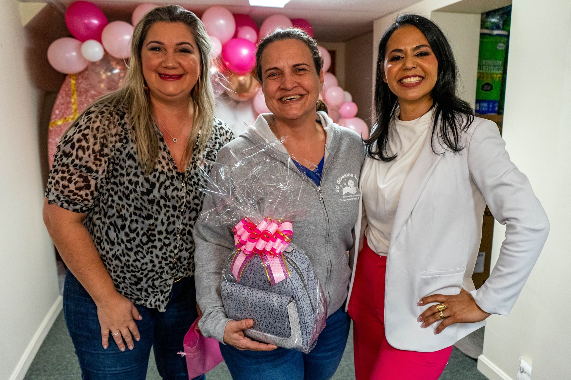 Three women smiling, posing with a gift basket; pink balloons in the background.