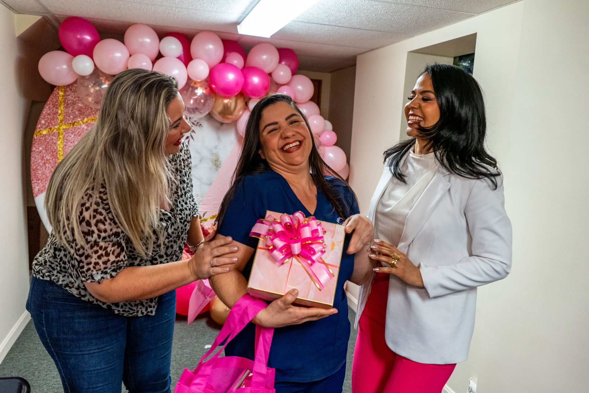 Three women celebrating. One holds a pink gift, others smile, pink balloons in background.
