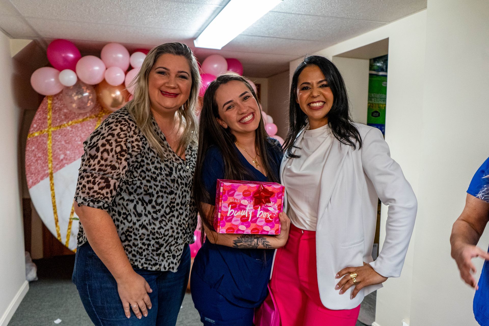 Three smiling women posing with a gift box, pink and gold balloons in the background.