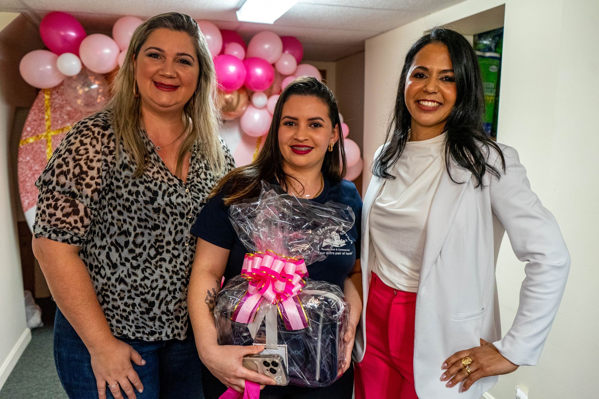 Three women pose, one holding gift basket. Pink and white balloon decor in background.