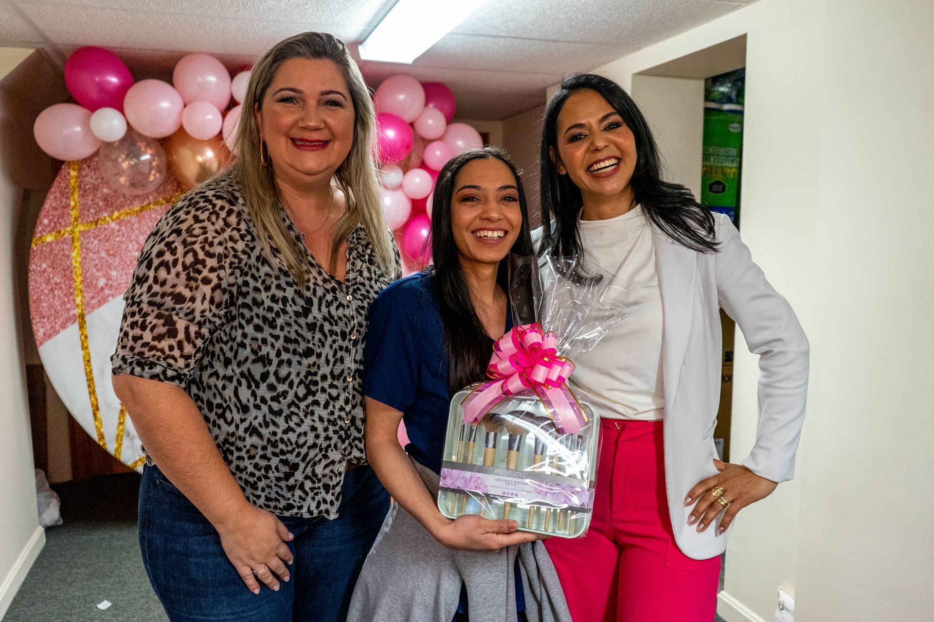 Three women pose with a gift basket, smiling. Pink and gold balloons in the background.