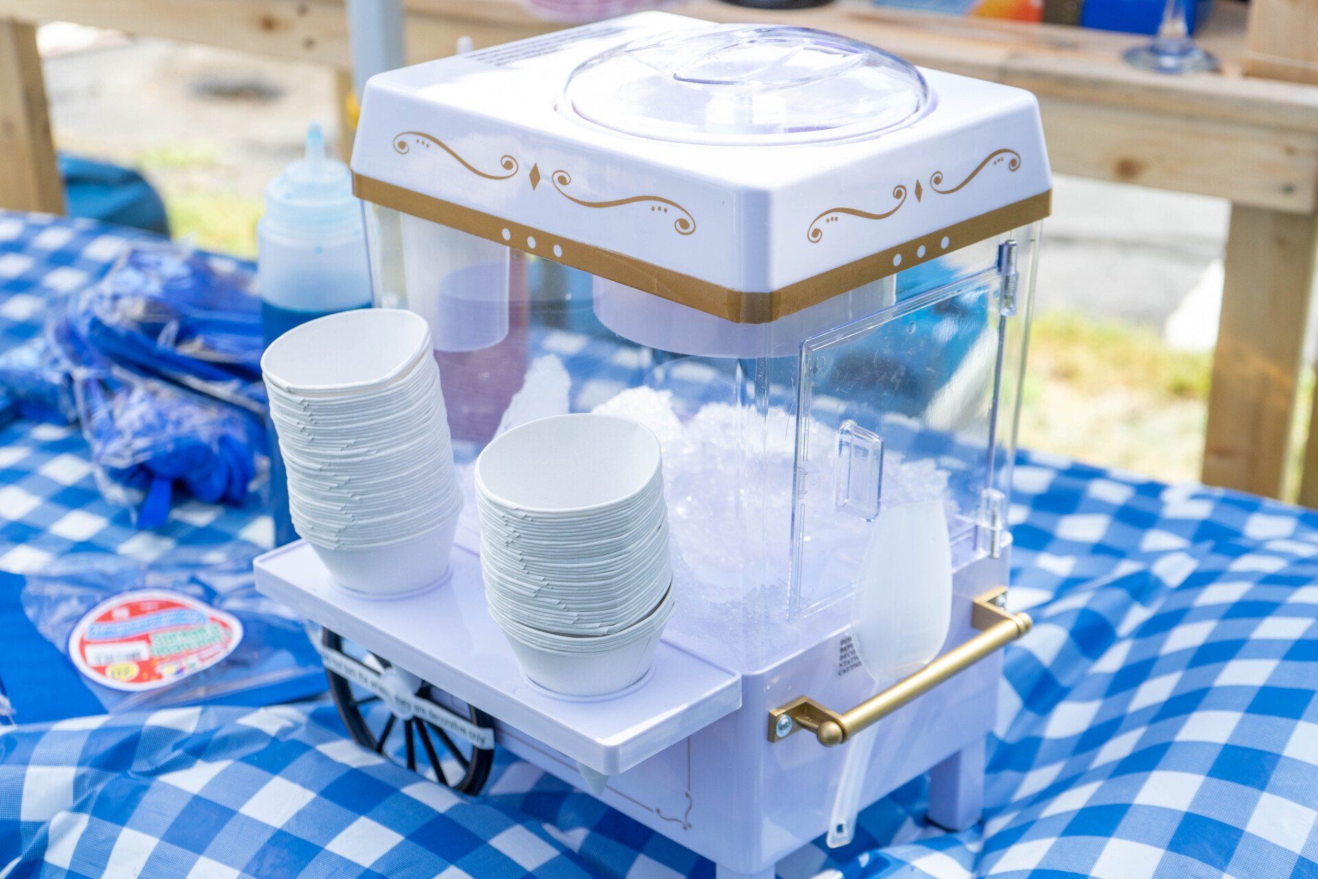 Snow cone machine with cups on a blue checkered tablecloth.