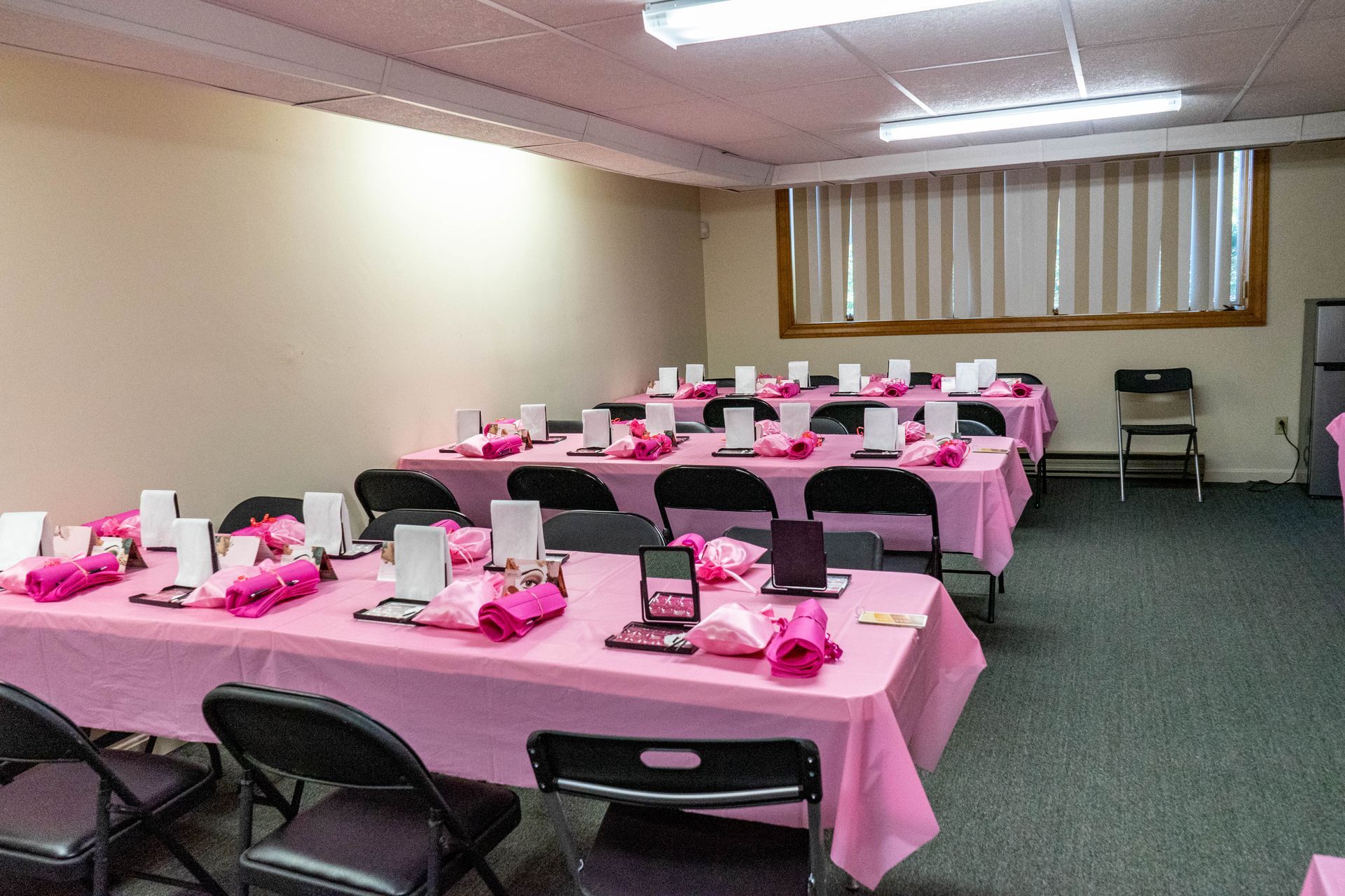 Pink-covered tables set for an event in a room with black chairs and a window.