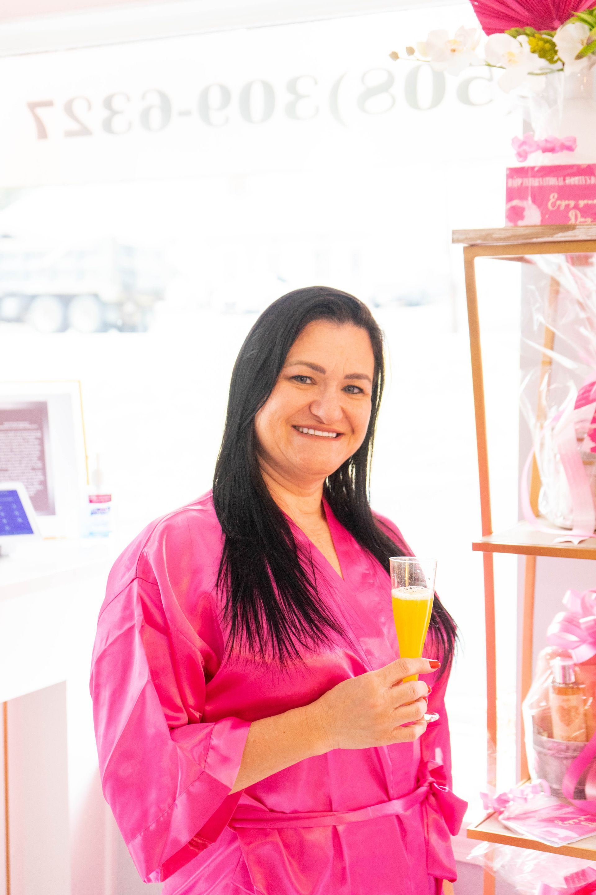 Woman in pink robe smiles, holding drink in bright room with shelf of pink items.