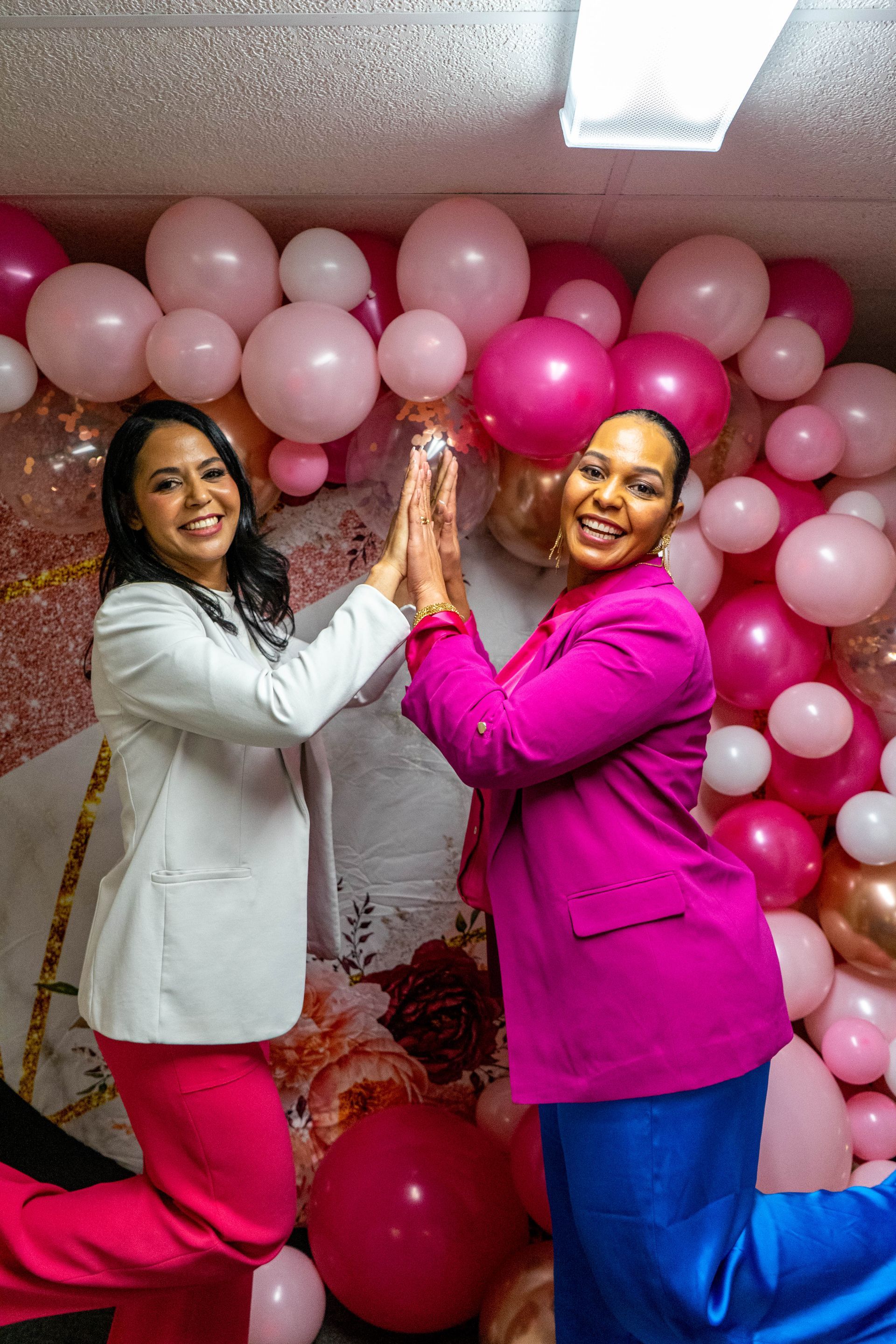 Two women high-fiving in front of pink balloons. One wears white blazer, pink pants. The other, a pink blazer, blue pants.