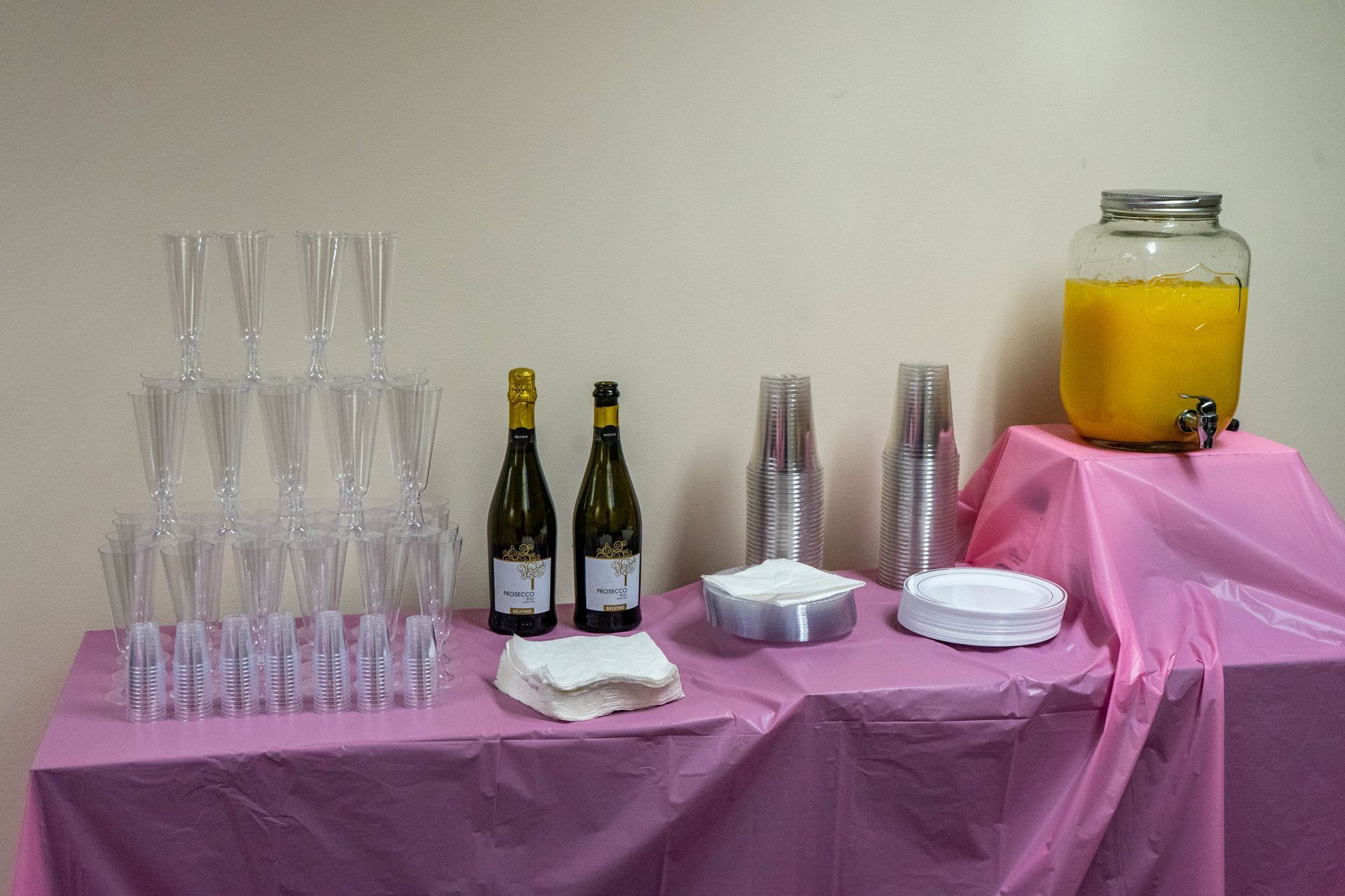 Pink-covered table with champagne flutes, bottles, drinks, cups, and plates for an event.