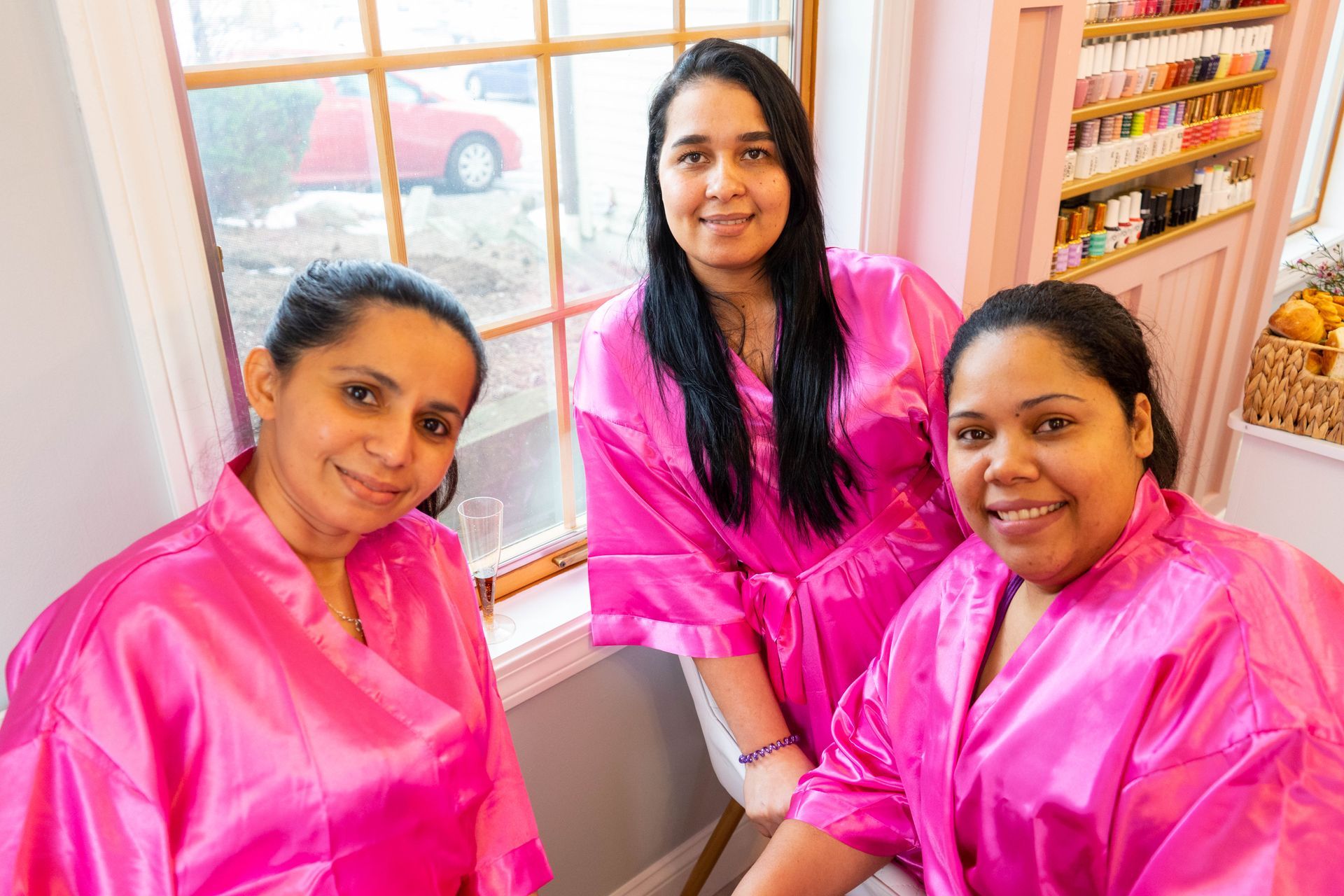 Three women in pink robes smiling by a window in a pink-themed room.