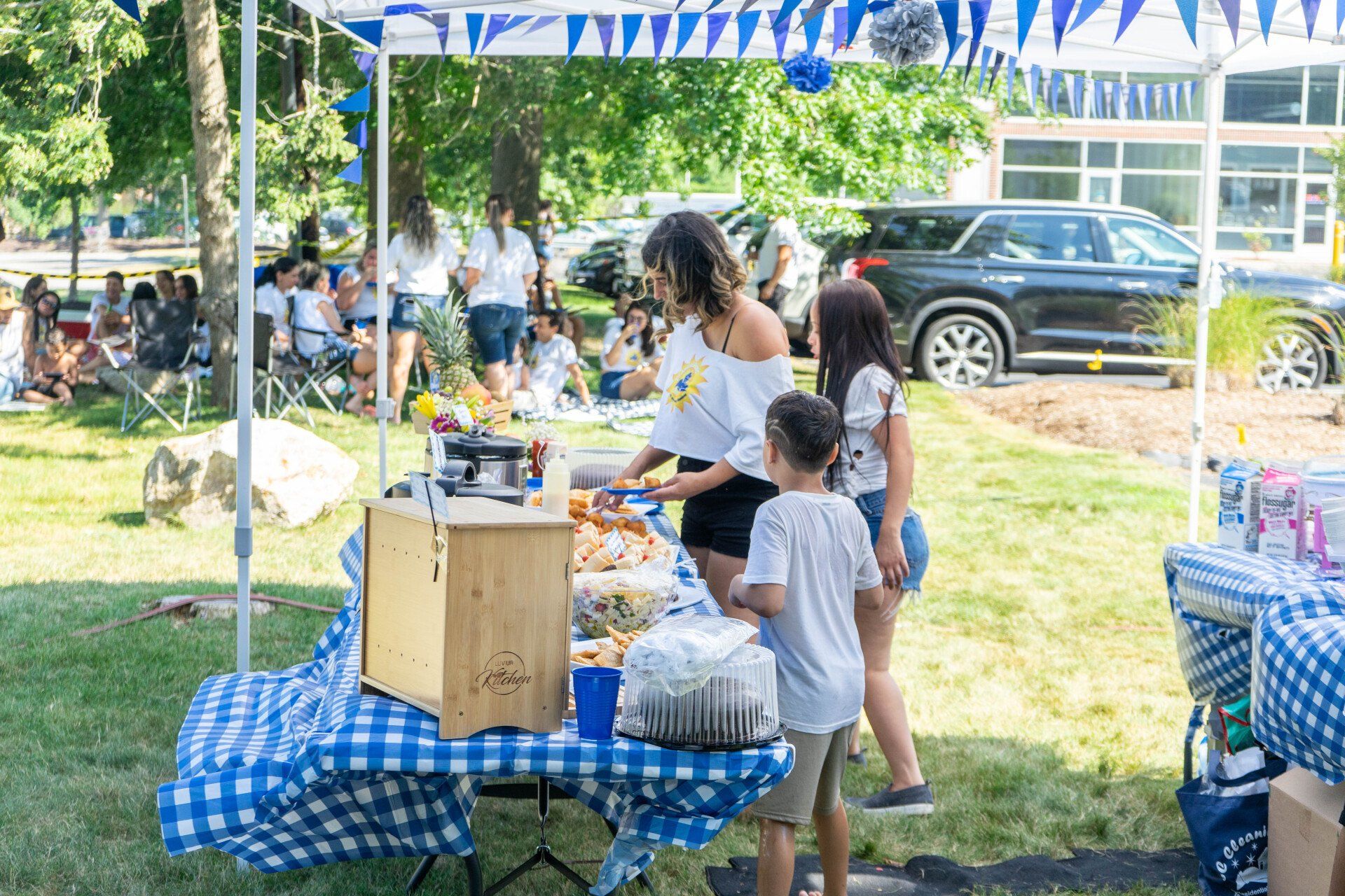 People at an outdoor event with a food table covered in blue and white checkered cloth.