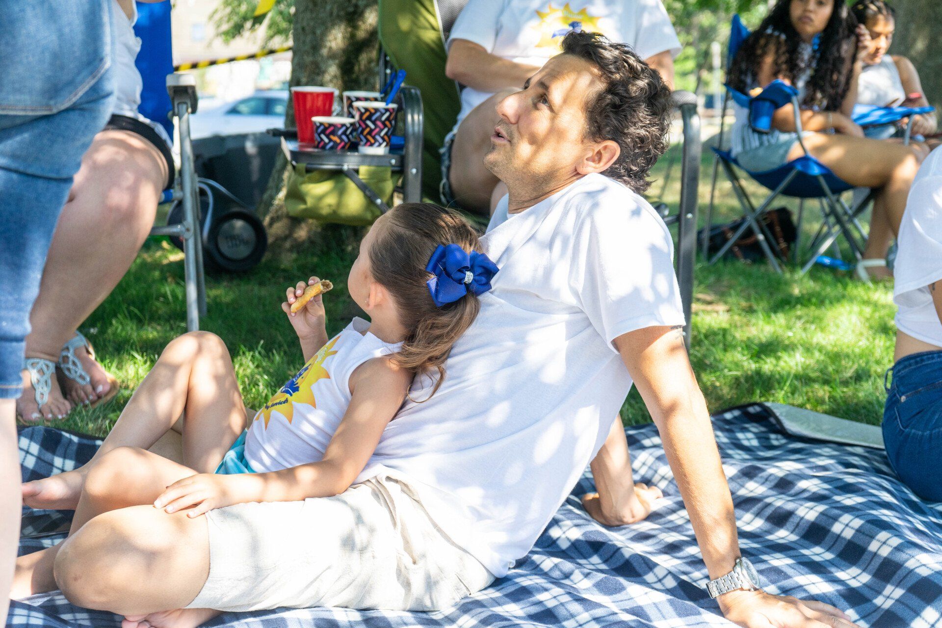 Father and daughter on a picnic blanket. The girl hugs the father who laughs. Blue sky, other people in background.