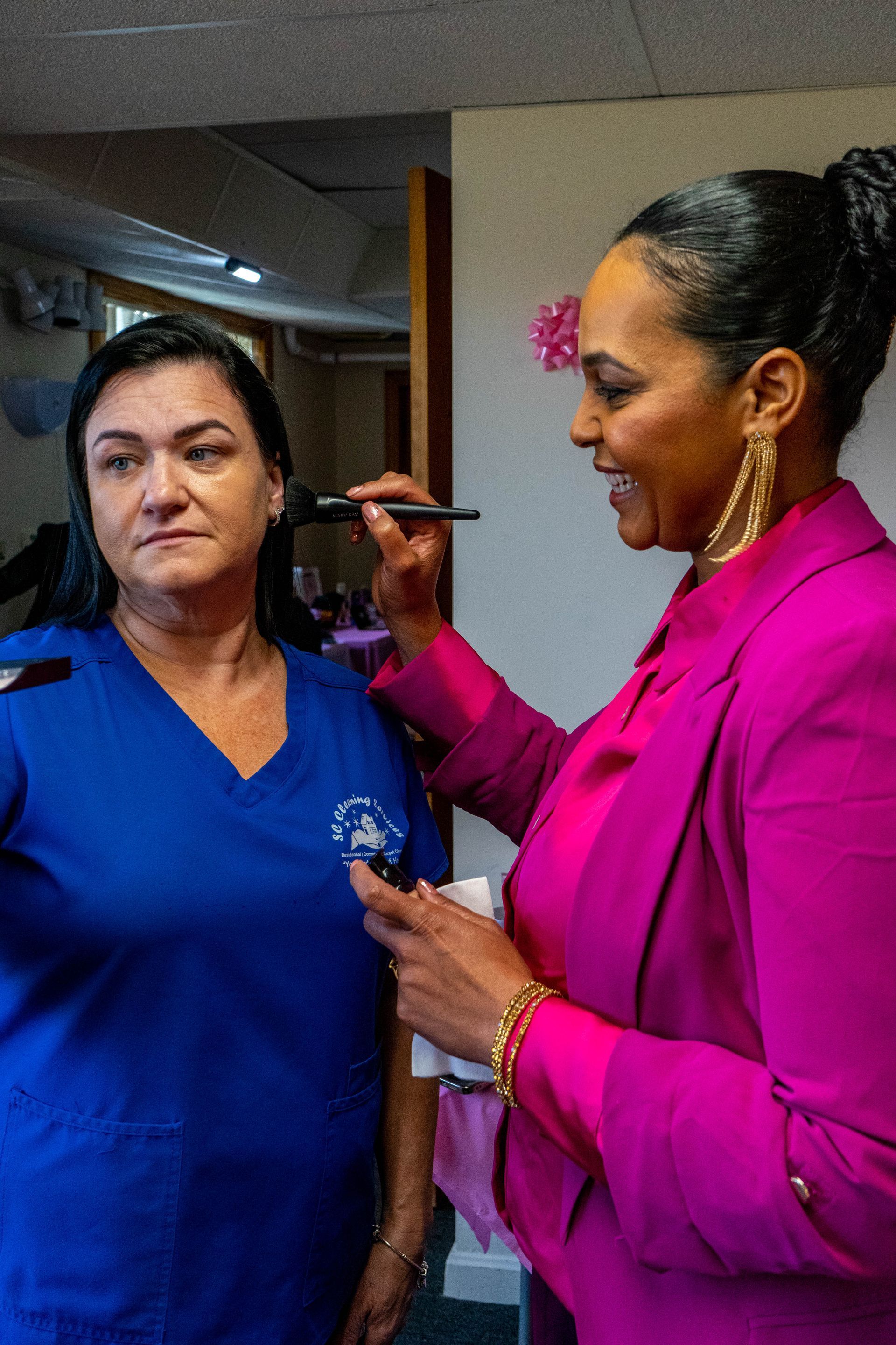 Woman in pink applies makeup to a woman in blue scrubs; both smiling in a brightly lit room.