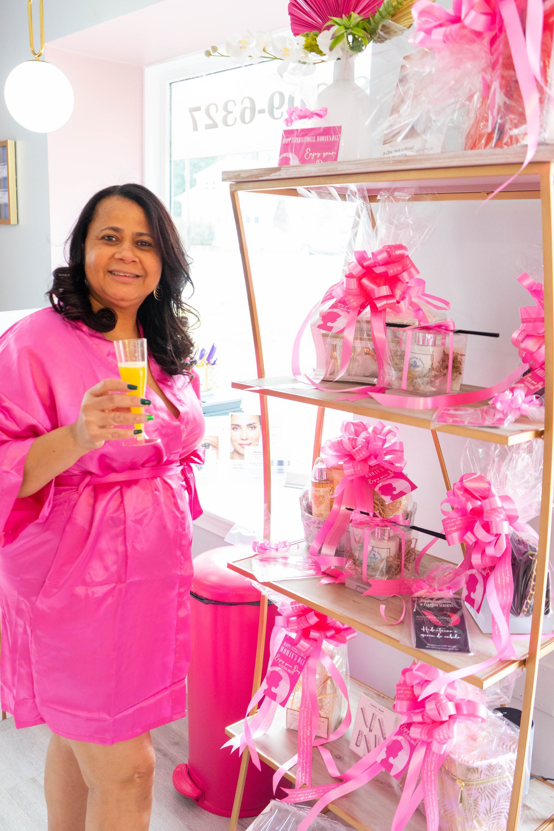Woman in pink robe holding drink, smiling, in a pink-themed shop with gift baskets and shelving.