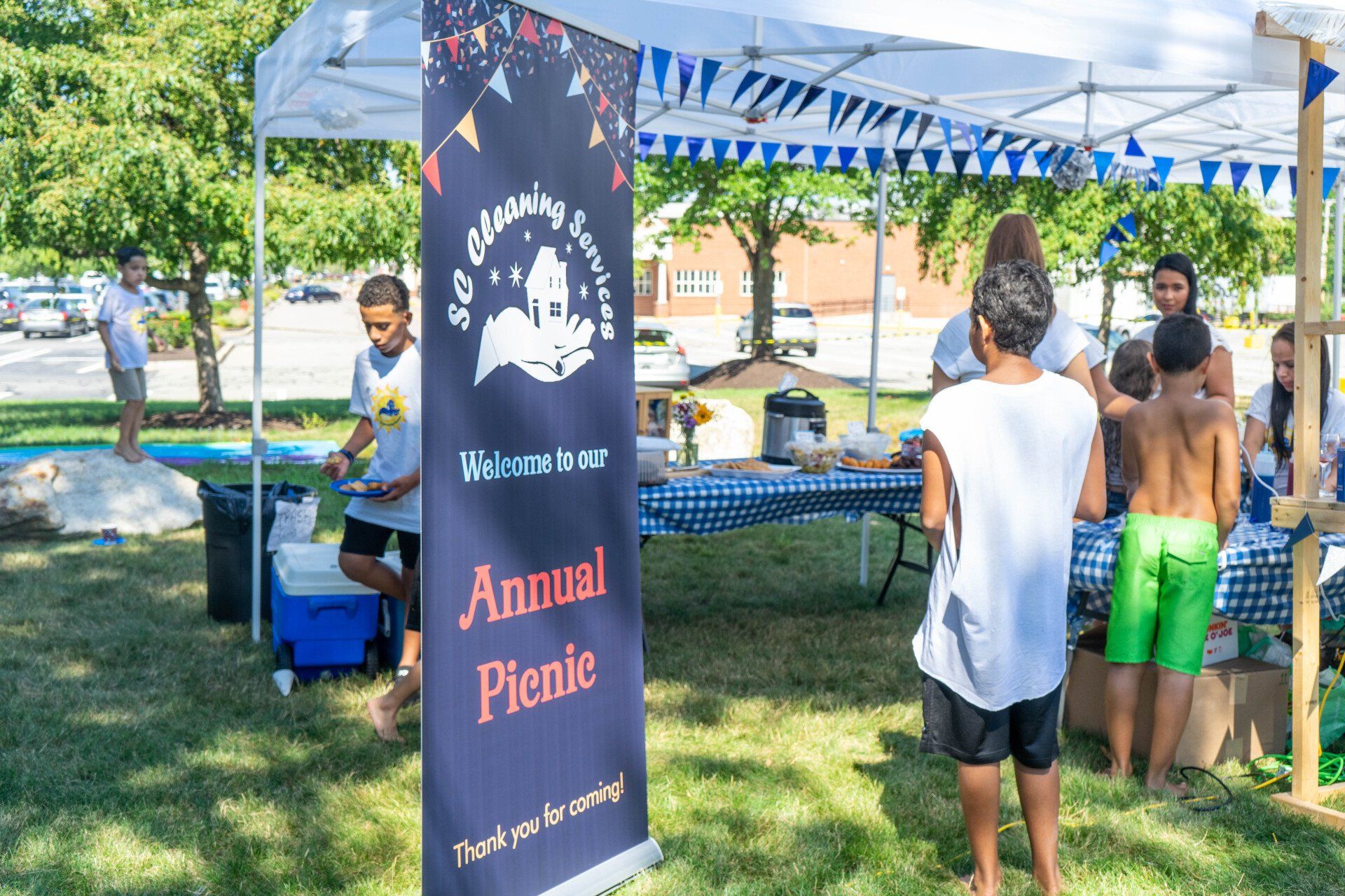 Annual picnic with people at a food table under a tent in a park. Banner, blue checkered tablecloth, sunny day.