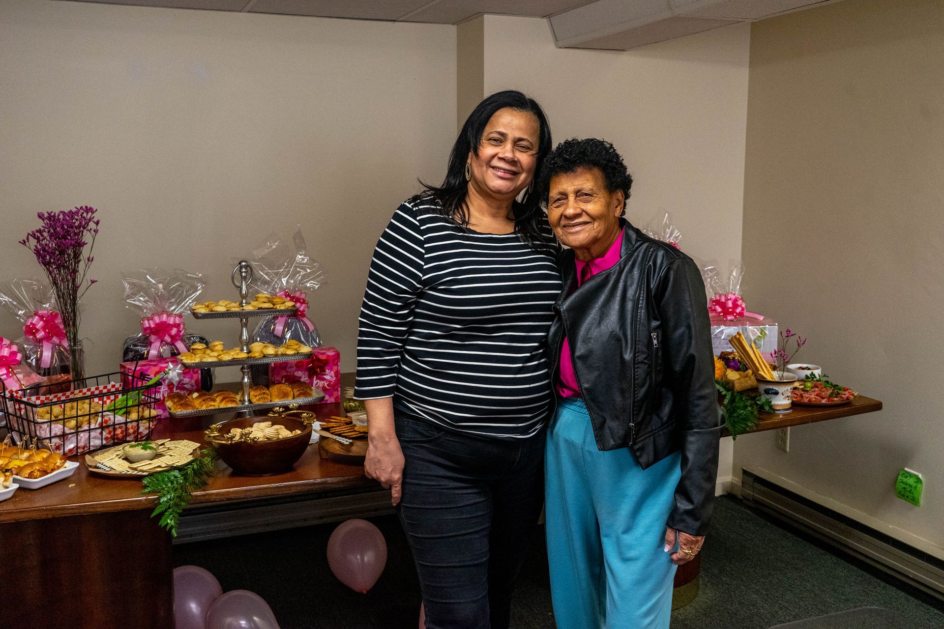 Two women pose near a table of food. One wears black and white stripes, the other wears turquoise pants and a leather jacket.
