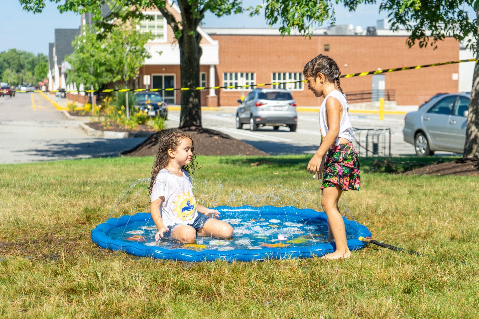 Two young girls playing in a blue inflatable pool on a grassy lawn; a building and cars are in the background.