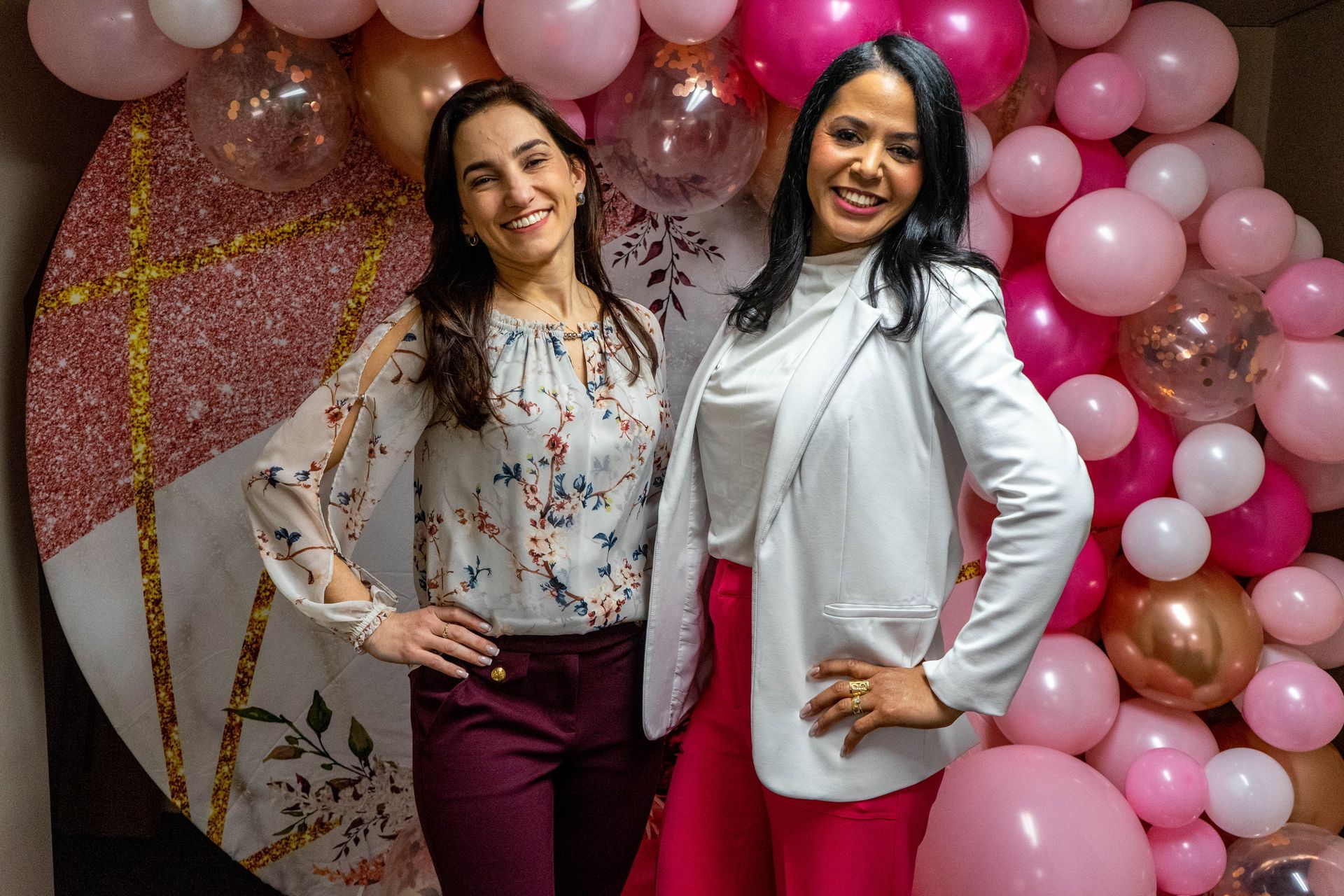 Two women smiling, posing in front of pink balloon backdrop, one wearing white blazer and pink pants.