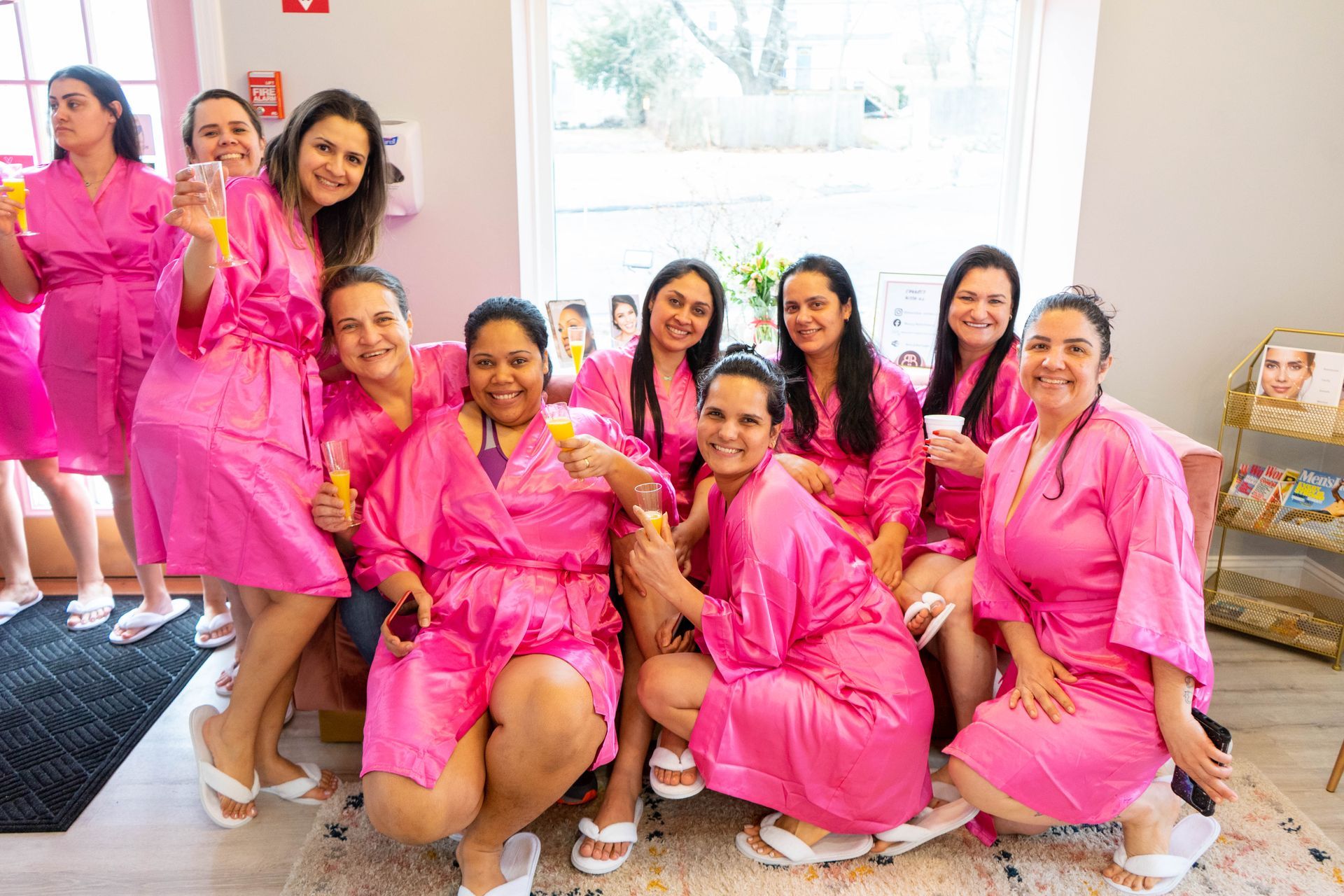 Group of women in pink robes, smiling and posing, holding drinks. They appear to be at a spa or salon.