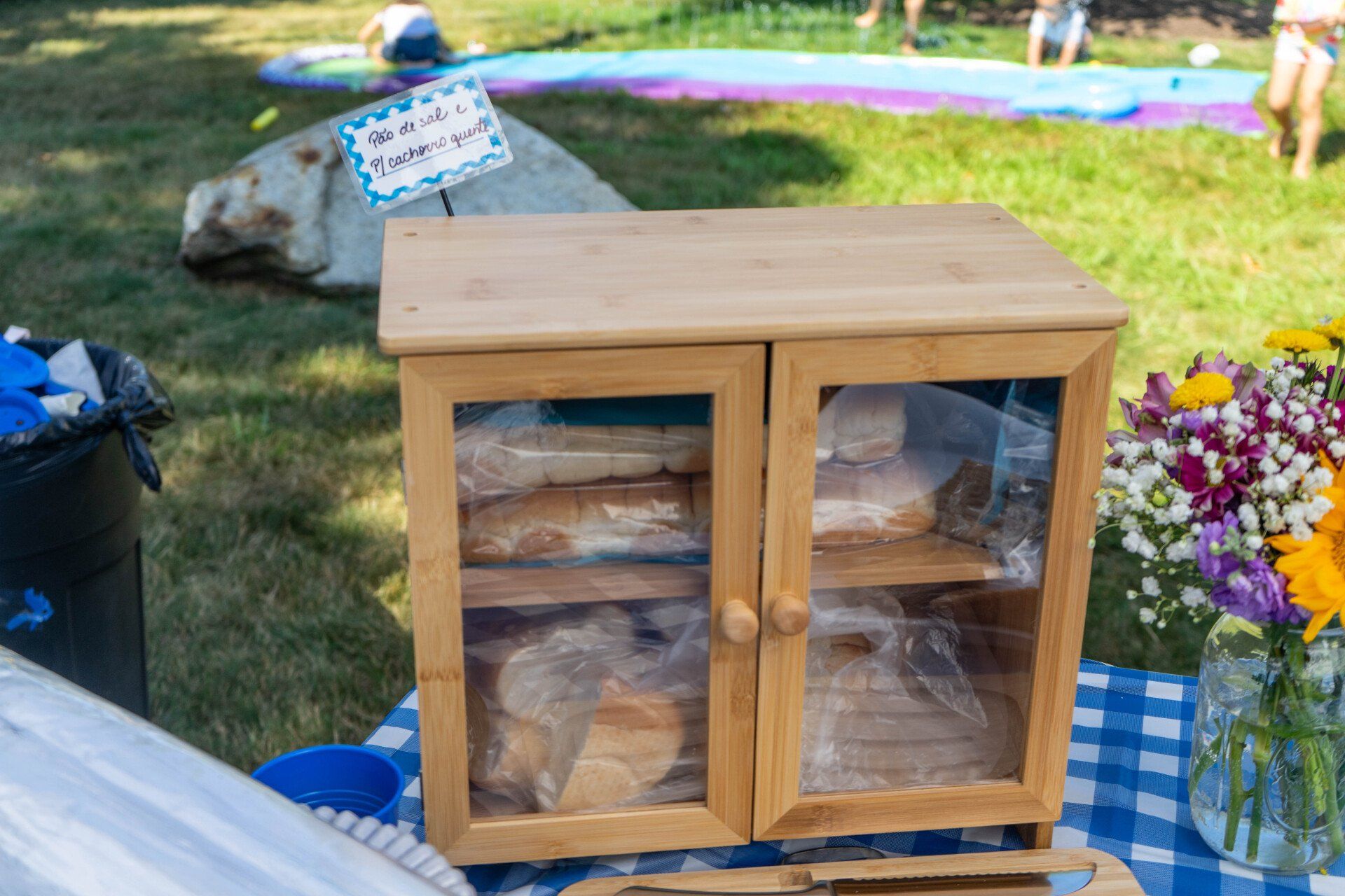 Wooden bread cabinet with loaves inside, outdoors on a blue checkered tablecloth.