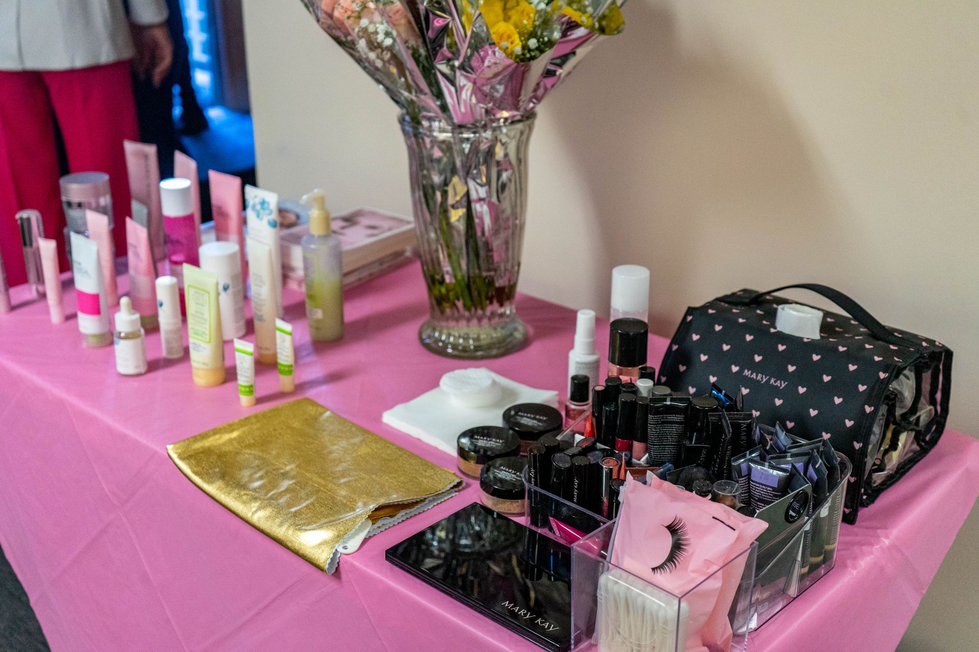 A table with cosmetics, including skincare products and makeup, arranged for display on a pink tablecloth.