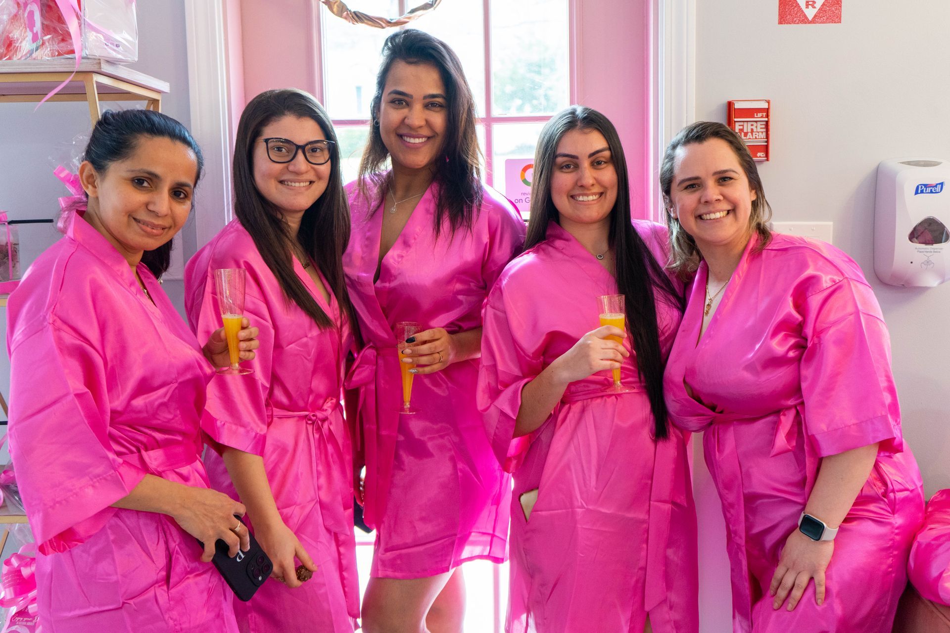 Five women in pink robes with drinks, smiling, posing indoors.