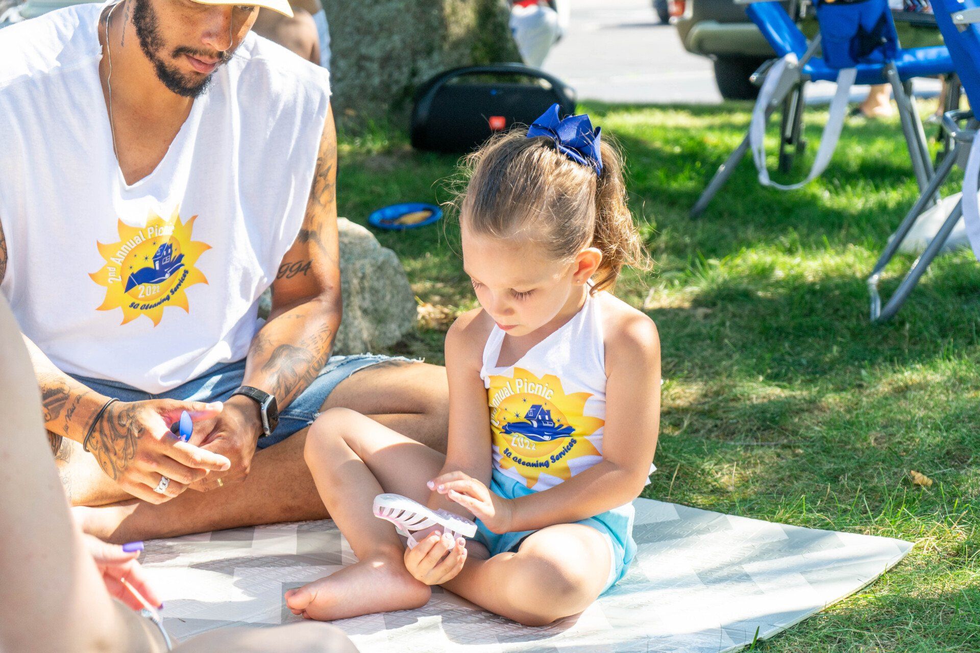 A young girl with a blue bow plays cards with an adult, sitting on a blanket outdoors. Both wear matching white shirts.