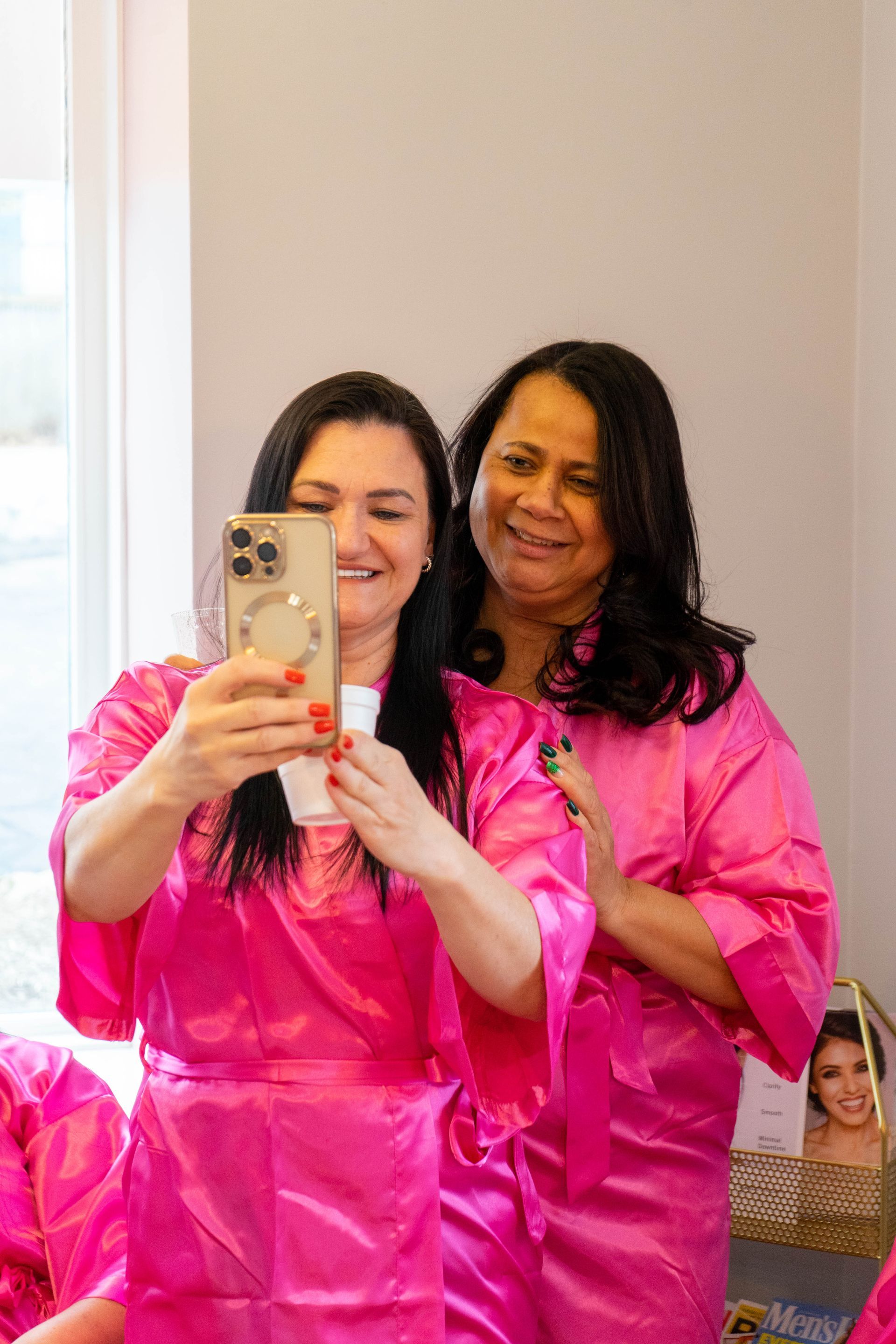 Two women in pink robes taking a selfie, smiling, in a brightly lit room.