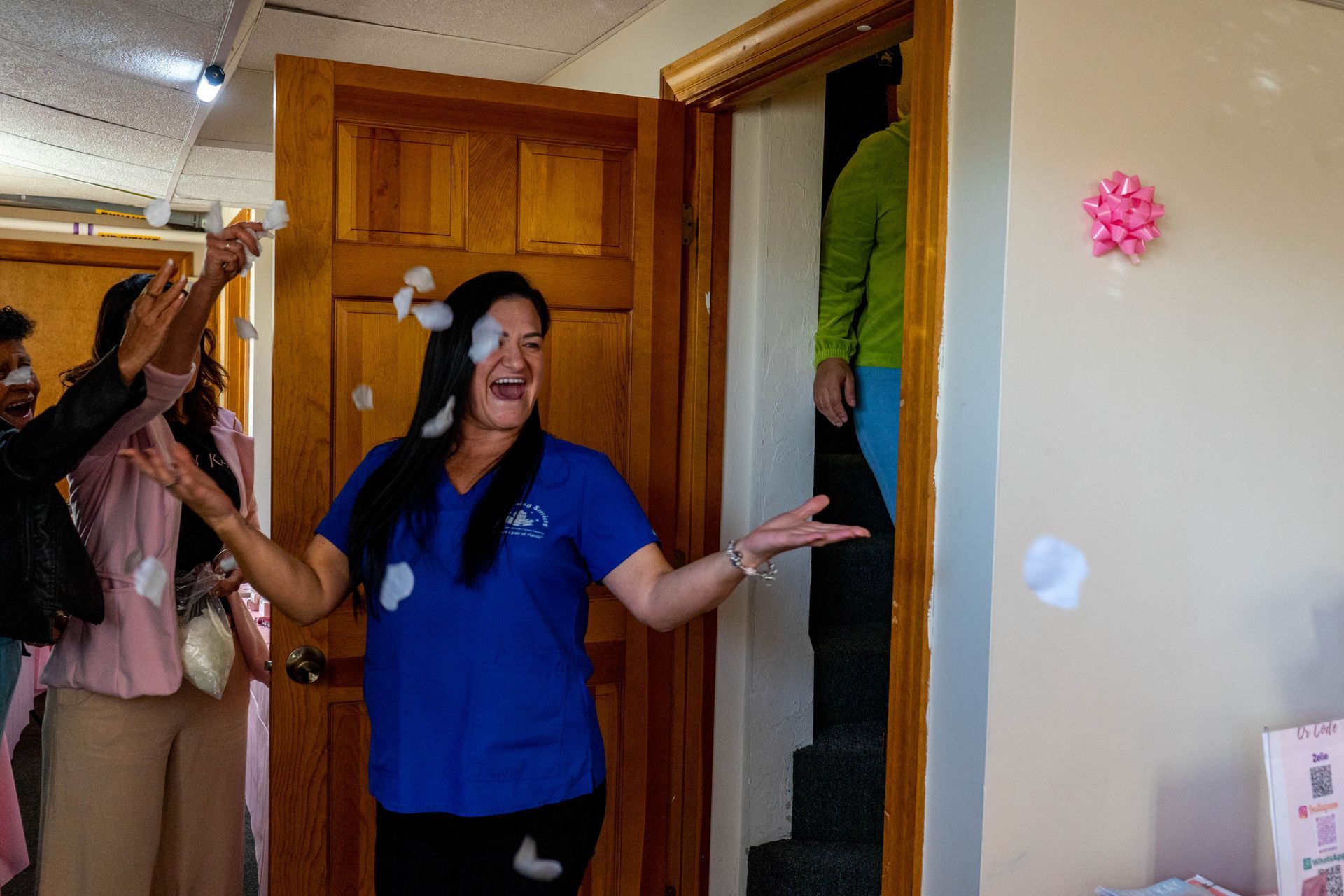 Woman in blue scrubs smiles as others toss confetti near an open doorway, pink bow on wall.