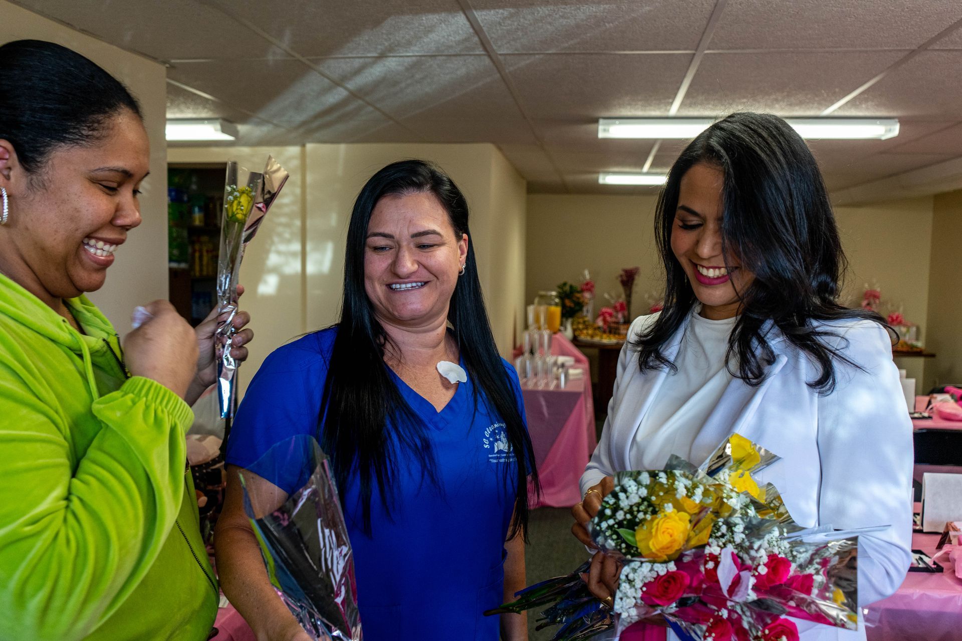 Three smiling women holding bouquets. One in green, one in blue scrubs, and one in a white jacket. Indoors.