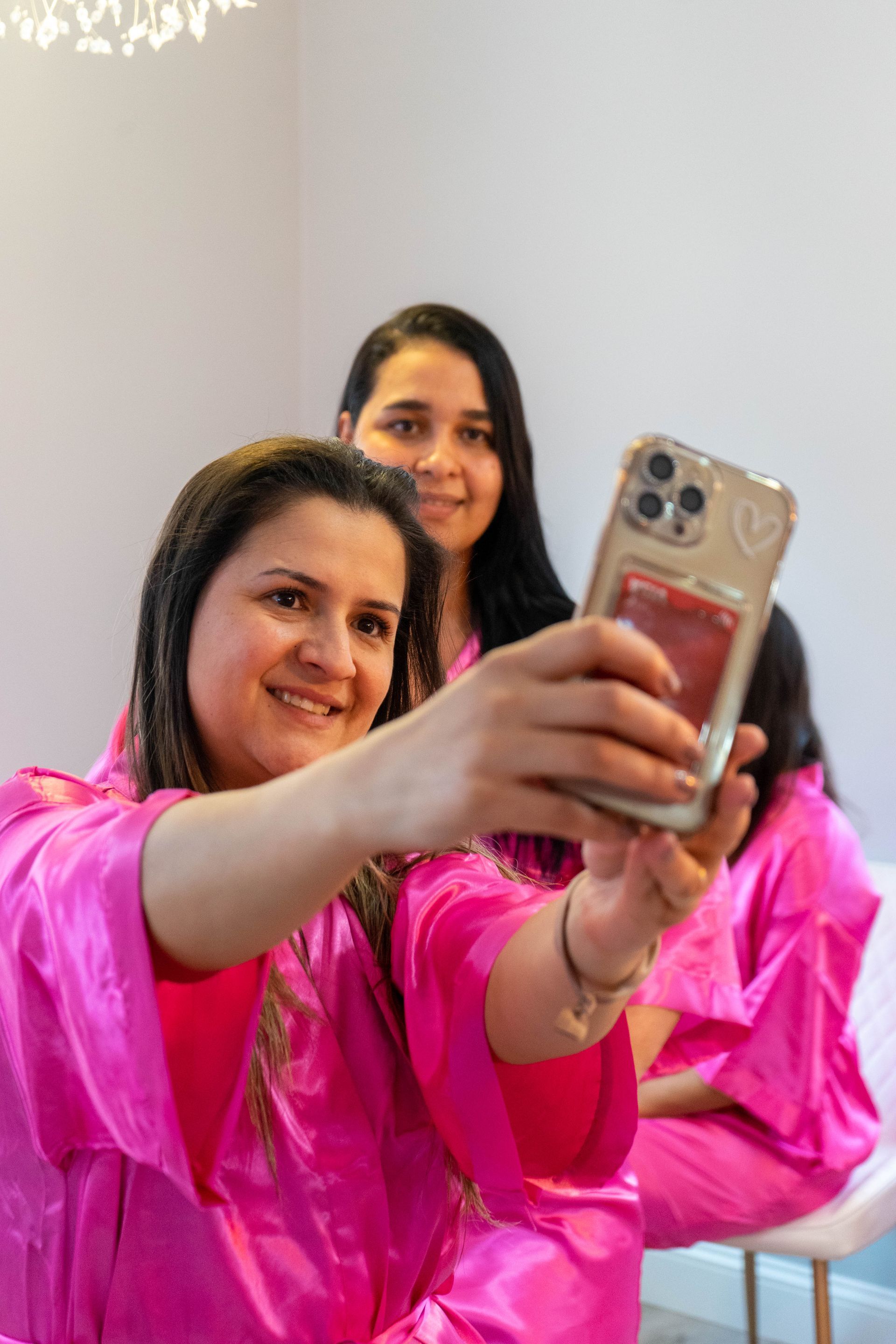 Three women in pink robes take a selfie, smiling, in a bright room.