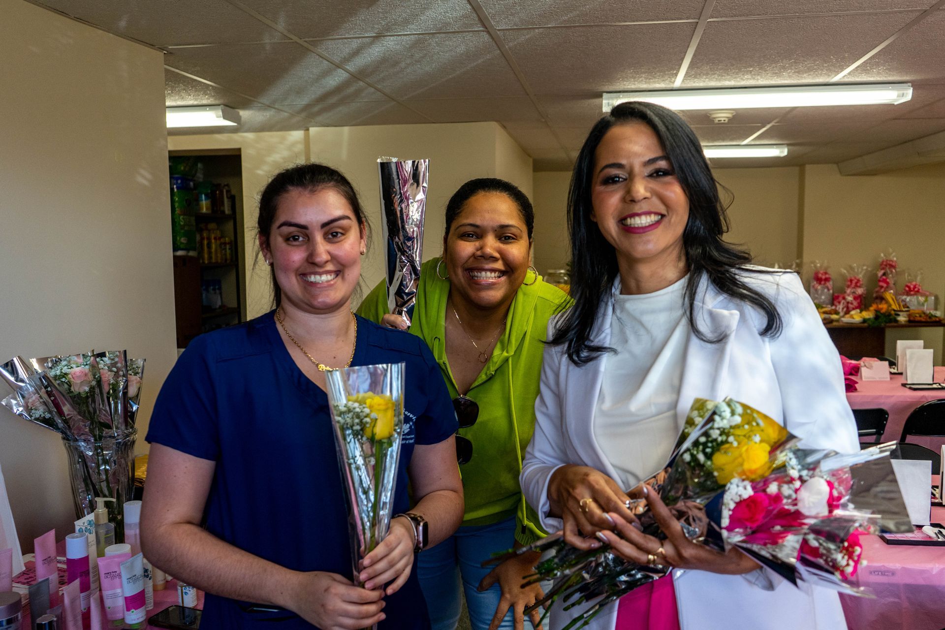 Three women smiling, holding flowers, in a room with gifts. One in white blazer.
