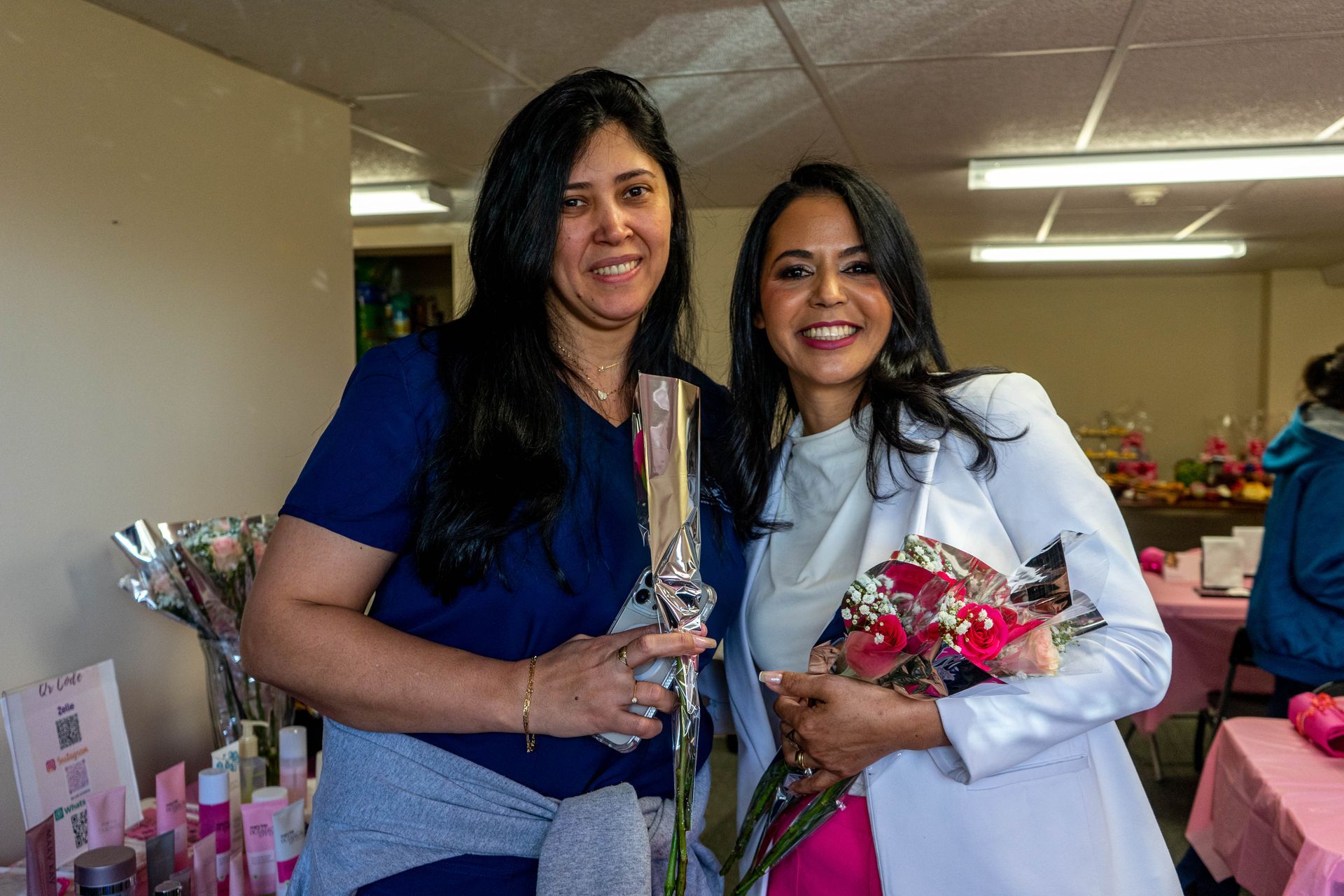 Two women smile, holding gifts and flowers at an event. One wears a white blazer.