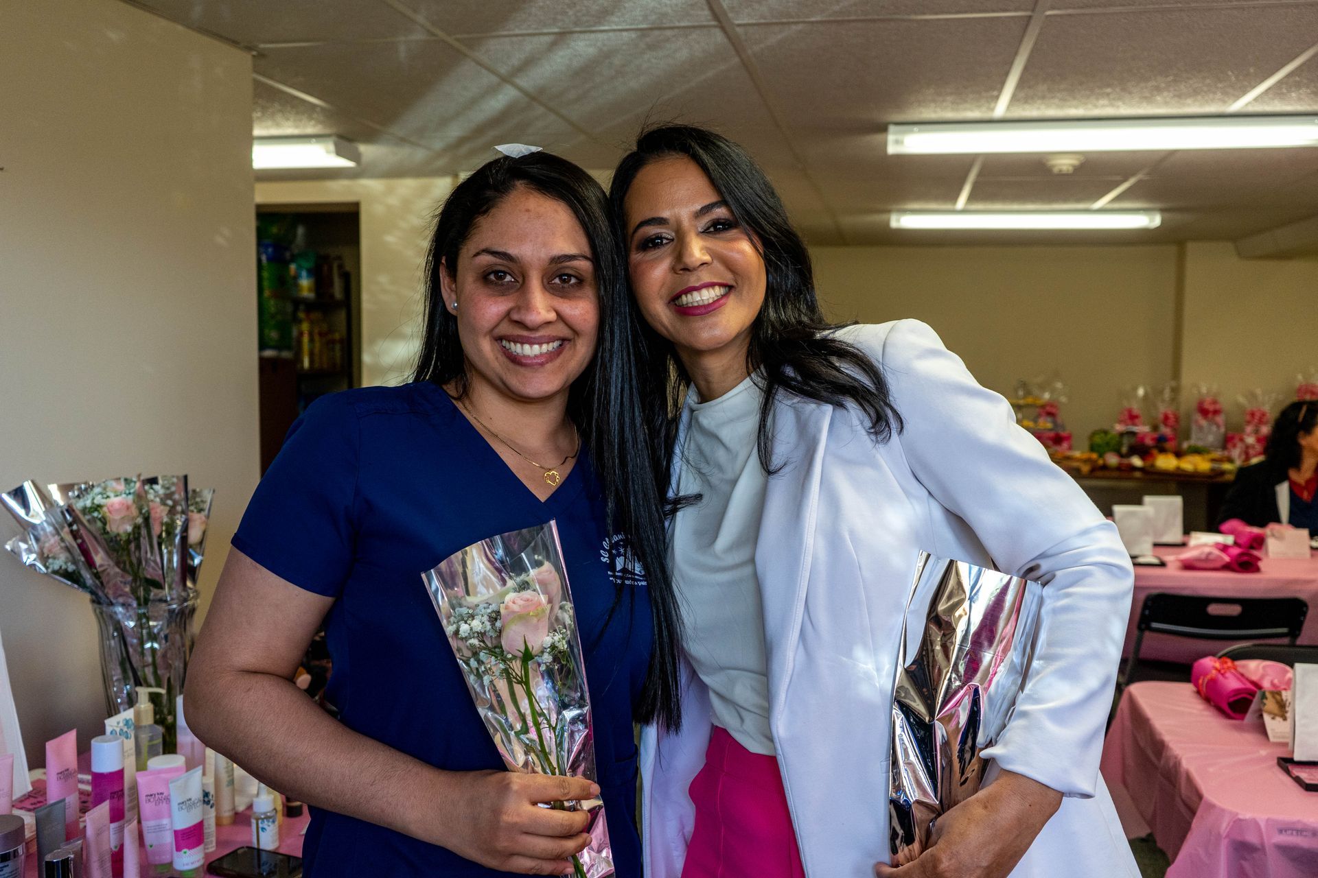 Two smiling women holding flowers, one in scrubs, one in a lab coat, at an event with pink decorations.