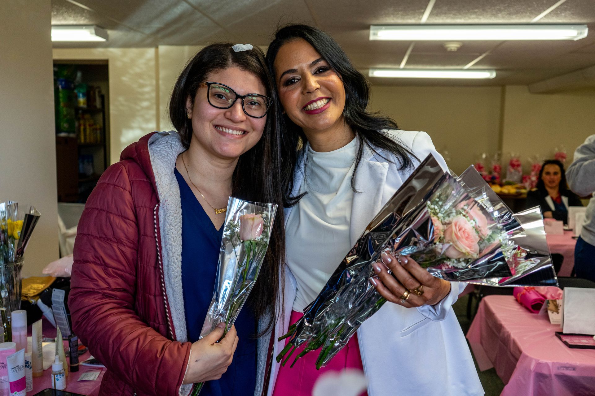 Two smiling women holding roses; one wears a white coat, the other a maroon jacket. They're inside, near a table.