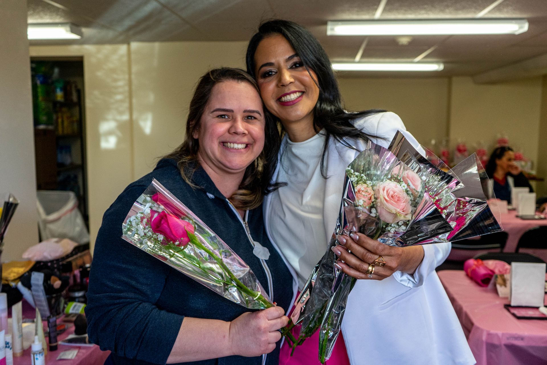 Two smiling women holding bouquets pose together at a pink-themed event.