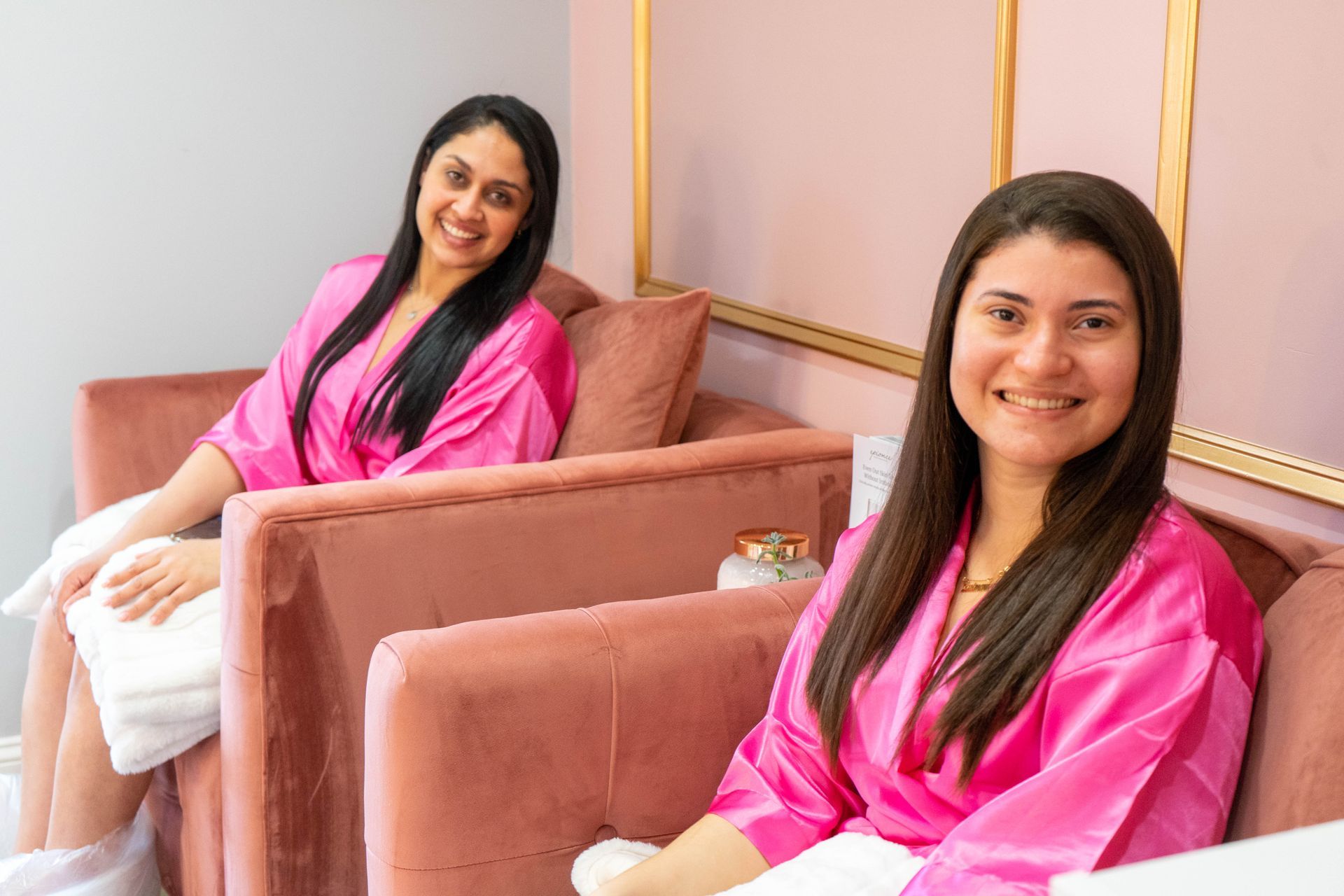 Two women in pink robes smile while getting pedicures in a pink salon.