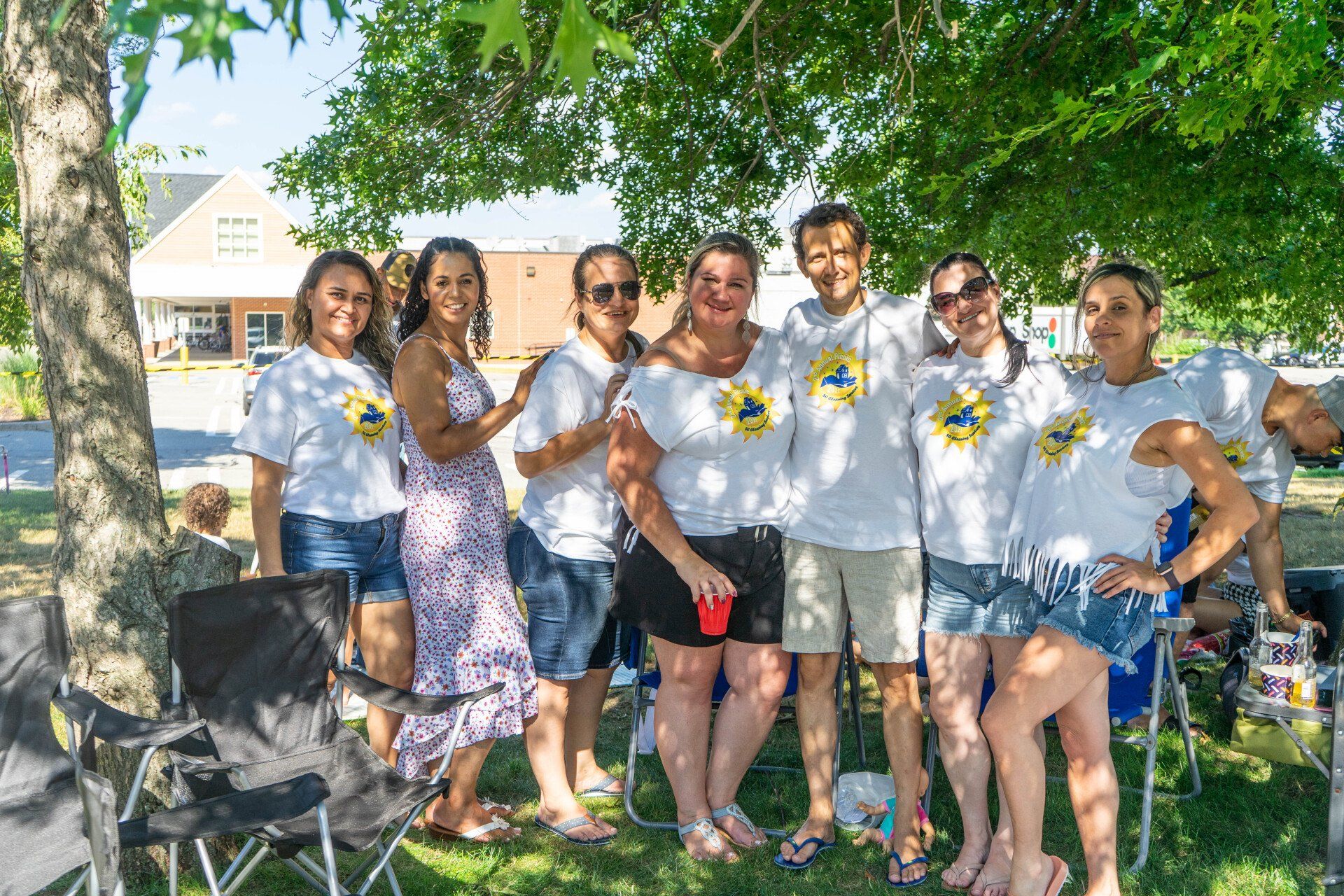 Group of friends smiling, posing outdoors under a tree. Some wearing matching white t-shirts.