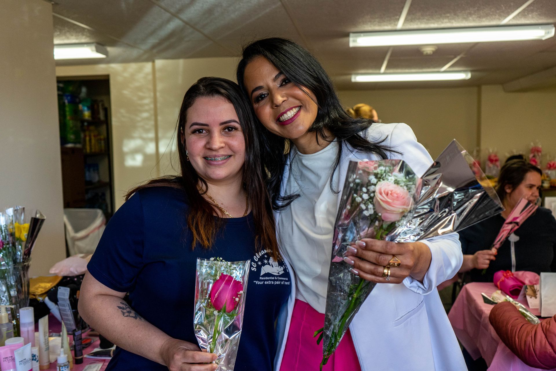 Two women smiling, holding pink roses. One in a lab coat, the other in a blue shirt. Setting possibly a healthcare facility.