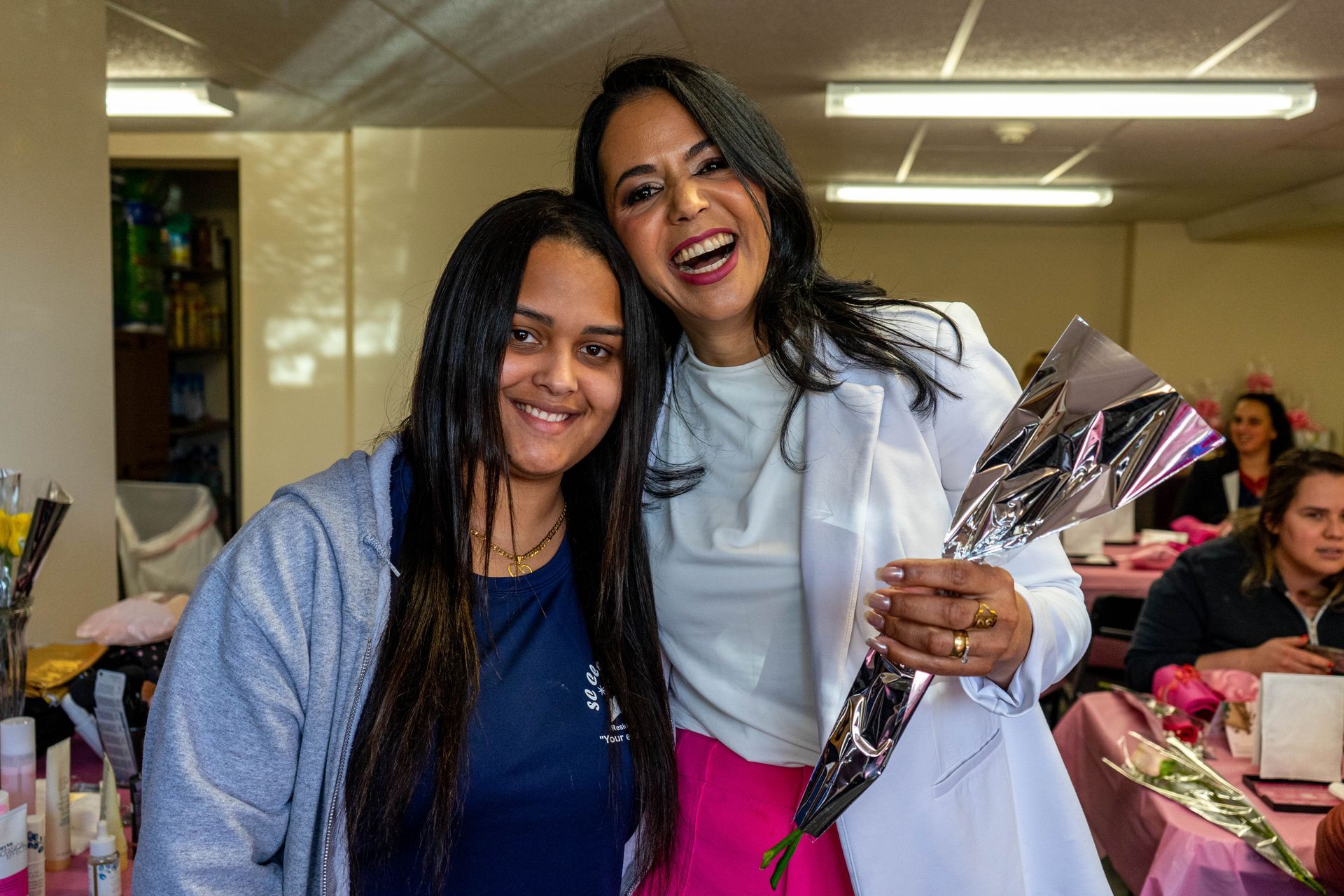 Two women smiling, embracing. One holds wrapped flowers, wearing a white jacket and pink pants. Other wears a grey hoodie.