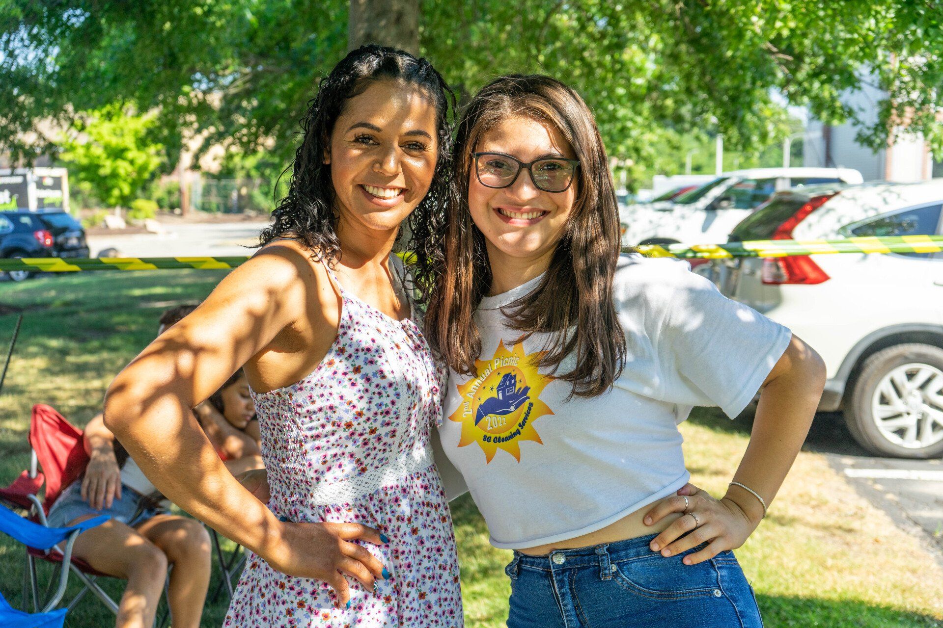 Two smiling women posing outside. One in a floral dress, the other in a white tee and jeans. Sunny, outdoor setting.