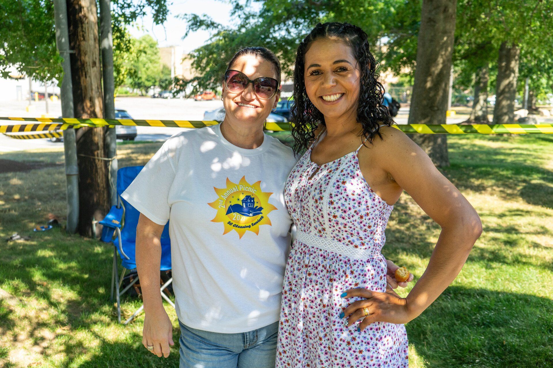 Two women smiling, one in a white floral dress, the other in a t-shirt, standing in a park on a sunny day.