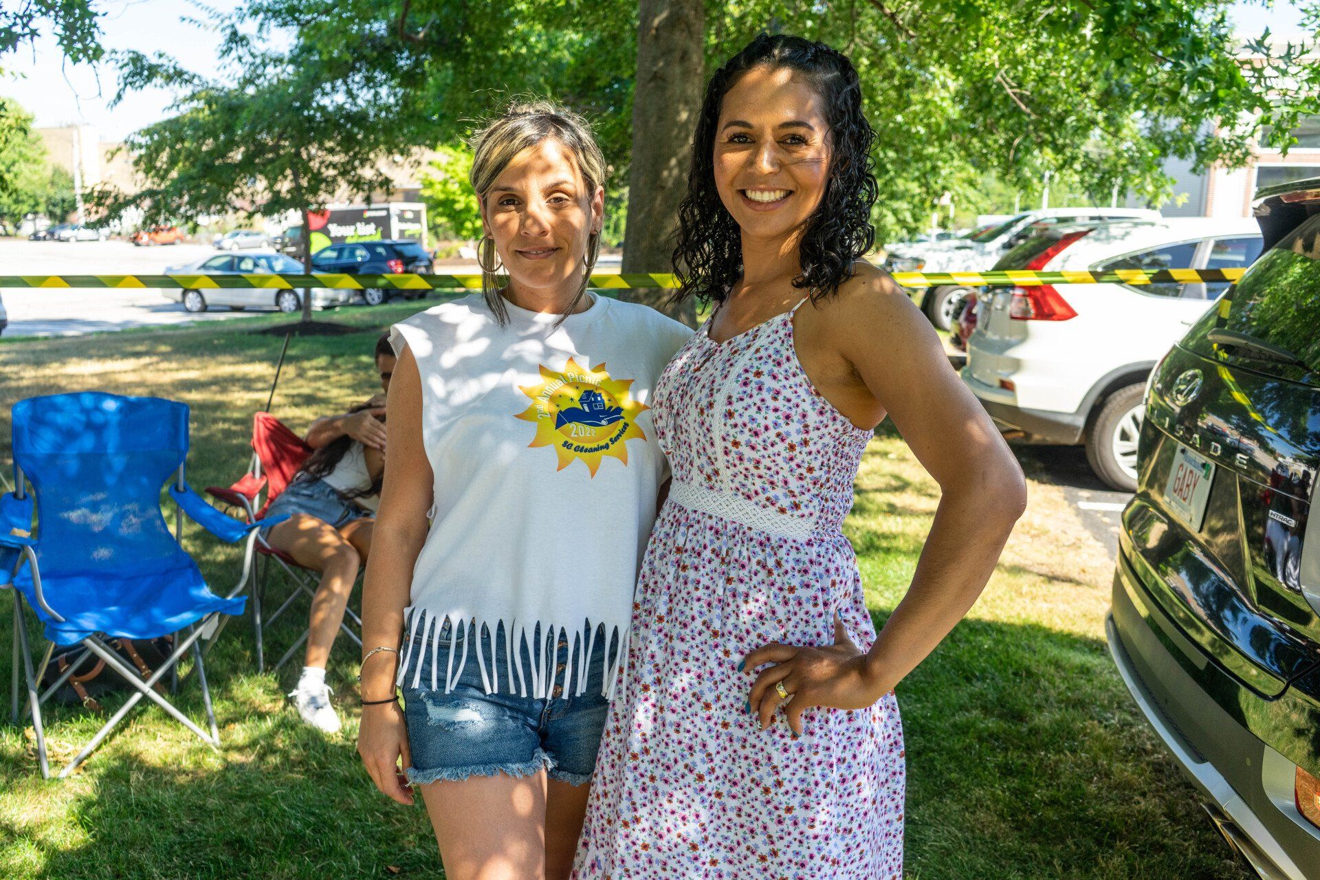 Two women smiling outdoors; one in a white fringe t-shirt and denim shorts, the other in a floral dress.