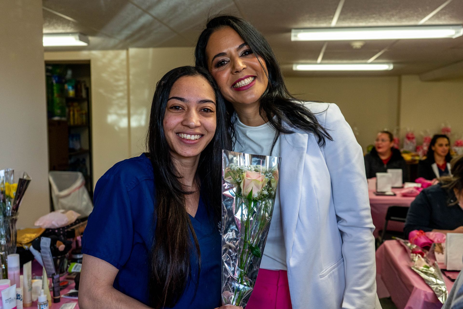 Two women smiling and embracing, one holding roses; setting appears to be an event with tables.