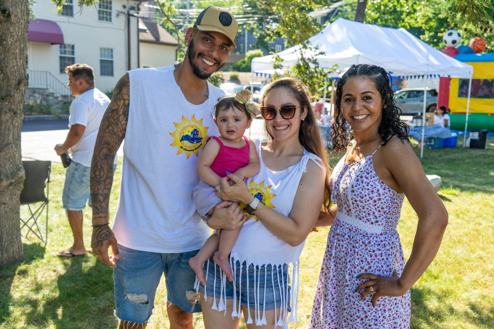 A family poses outdoors at an event; a man, woman holding baby, and another woman smile in front of a bouncy castle.