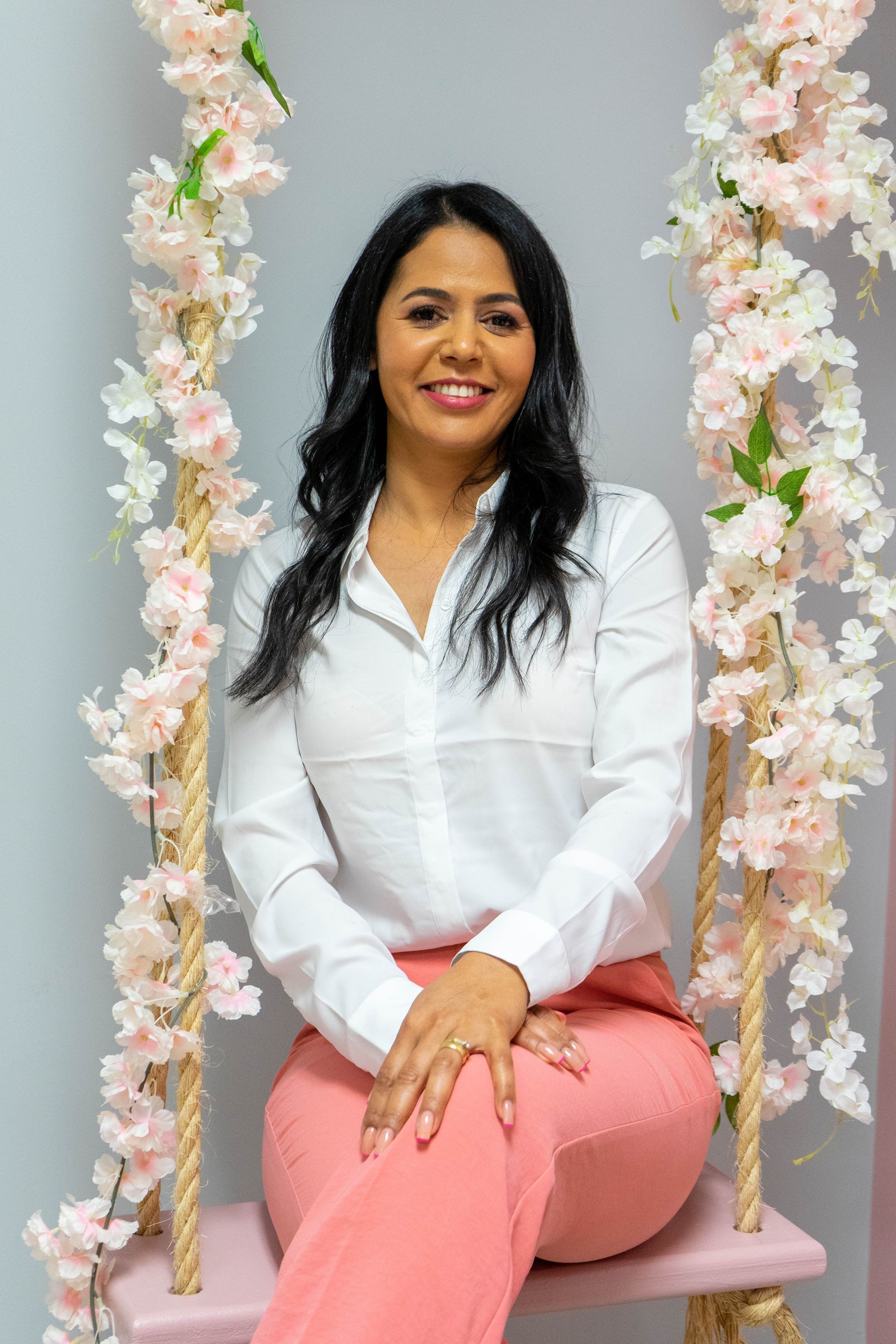 Woman seated on pink swing, wearing white shirt and pink pants, smiling. Pink floral decor in background.