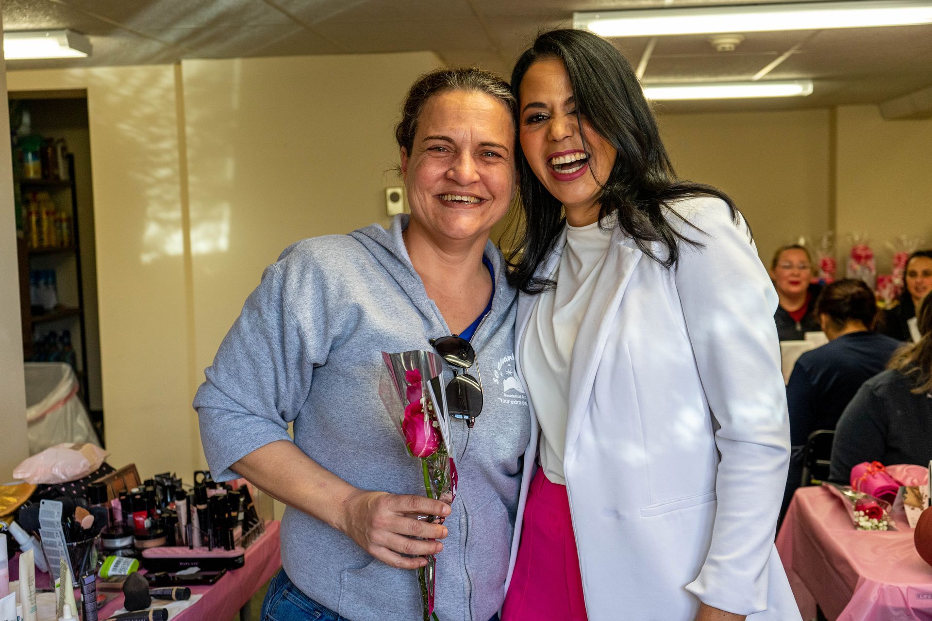 Two smiling women pose, one holding a rose. One wears a grey hoodie, the other a white jacket, both indoors.