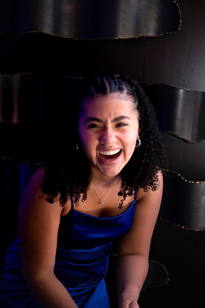 Woman with dark curly hair laughs widely, wearing a blue dress, against a dark textured background.